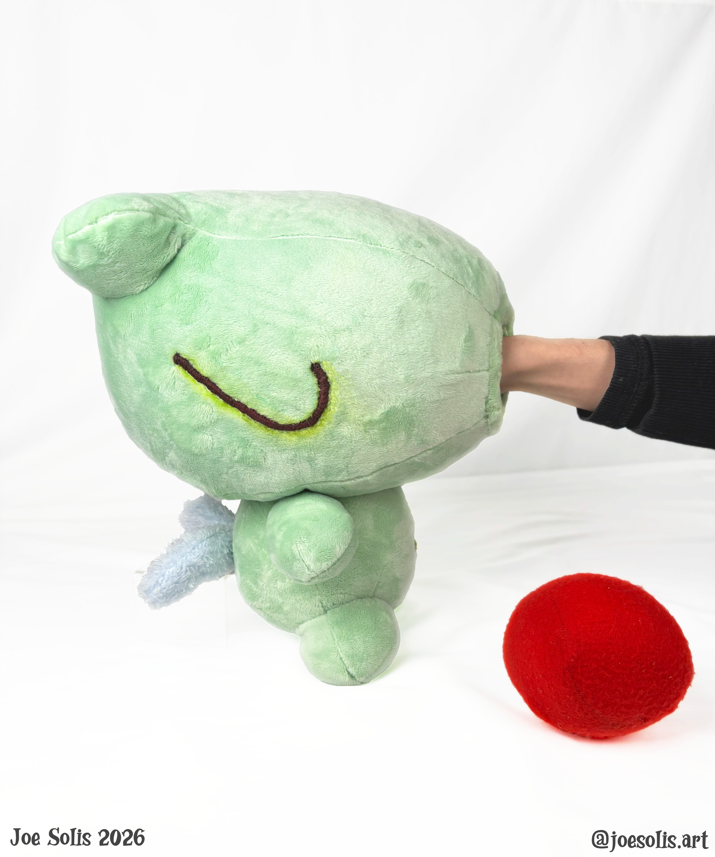 A green plush dinosaur with embroidered eyes and a smiling face, attached to a person's arm, with a red felt ball on the white surface below, and the text "Joe Solis 2026" and "@joasolis.art" at the bottom corners.