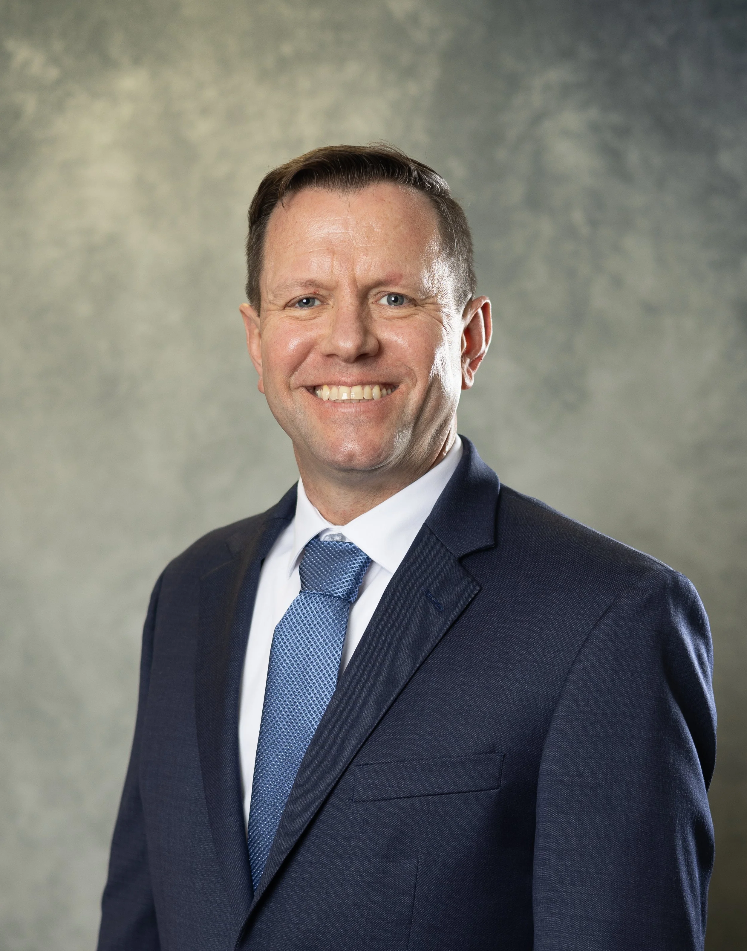 A smiling man in a navy suit, white shirt, and blue tie, standing against a textured gray background.