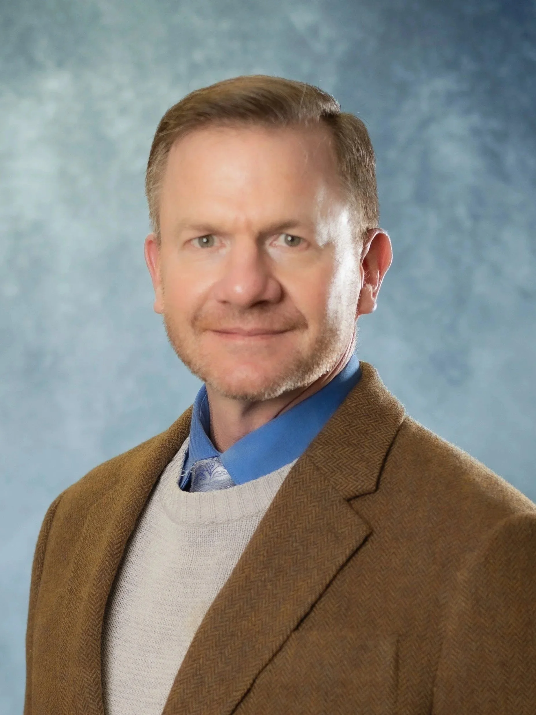 A professional headshot of a middle-aged man with fair skin, light brown hair, and a beard, dressed in a brown blazer, light blue shirt, and light-colored sweater, against a blue textured background.