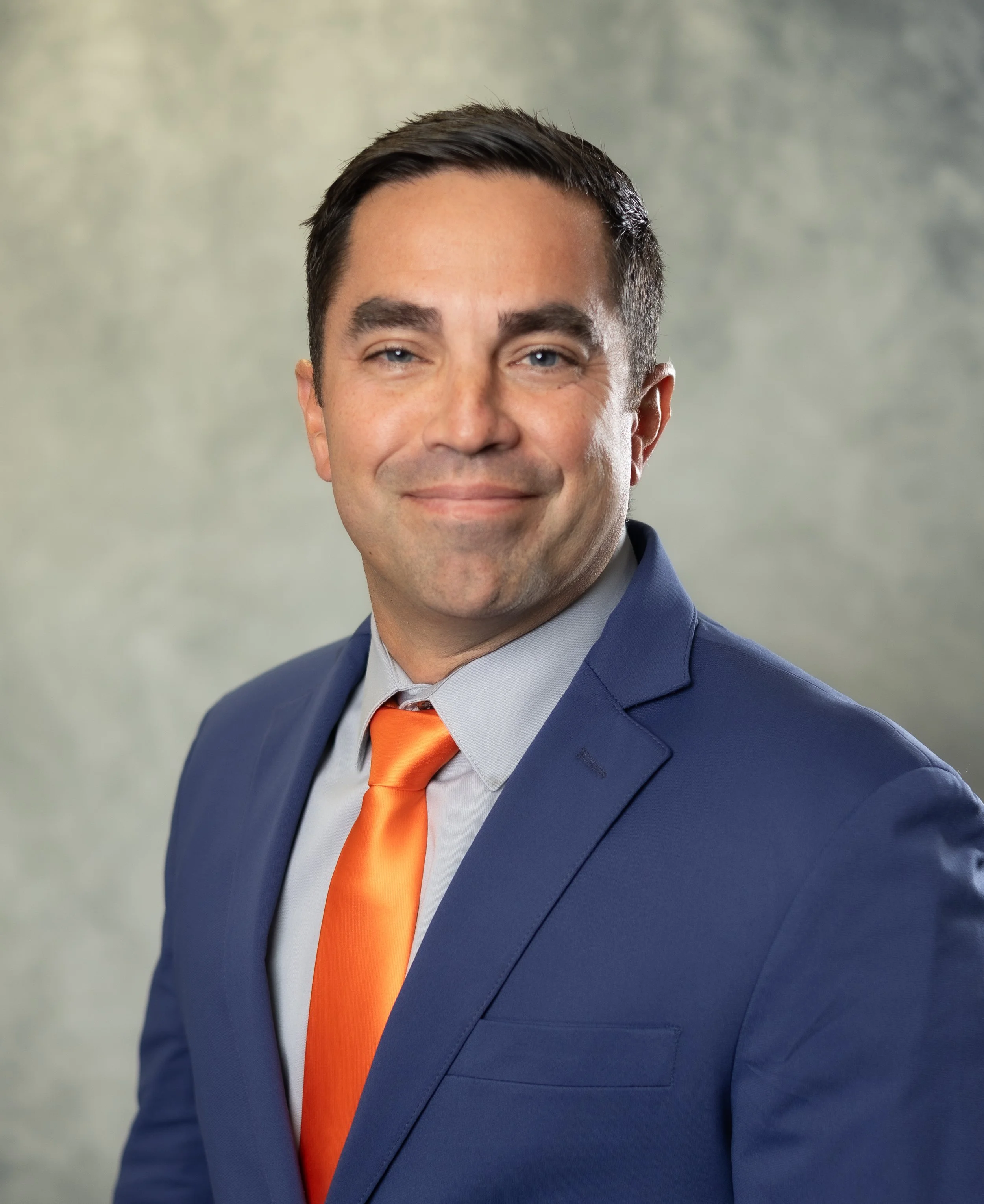 Professional headshot of a man in a blue suit, light blue shirt, and orange tie, smiling at the camera with a blurred gray and green background.