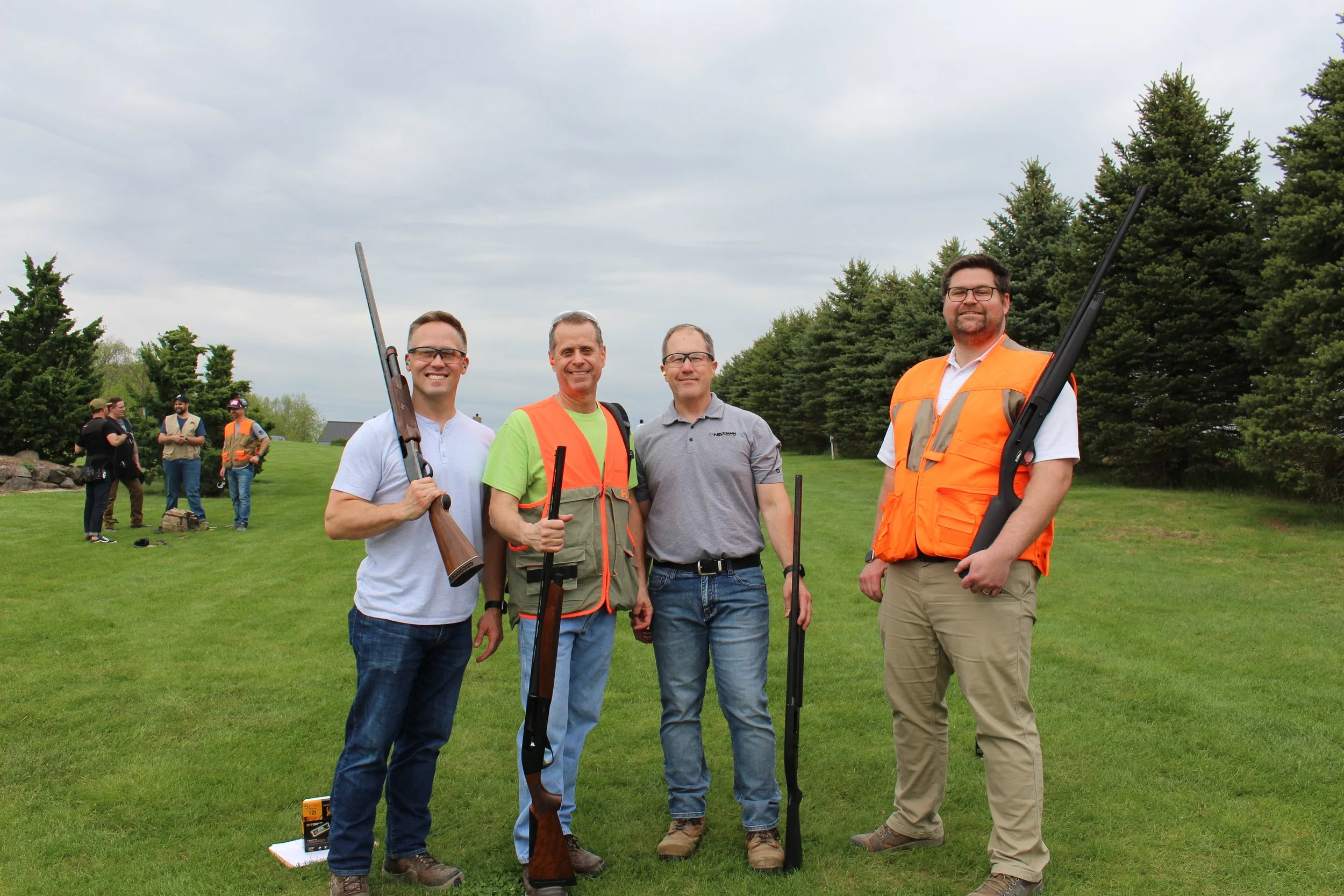 Four men standing on a grassy field holding shotguns, with others in the background, trees, and a cloudy sky.
