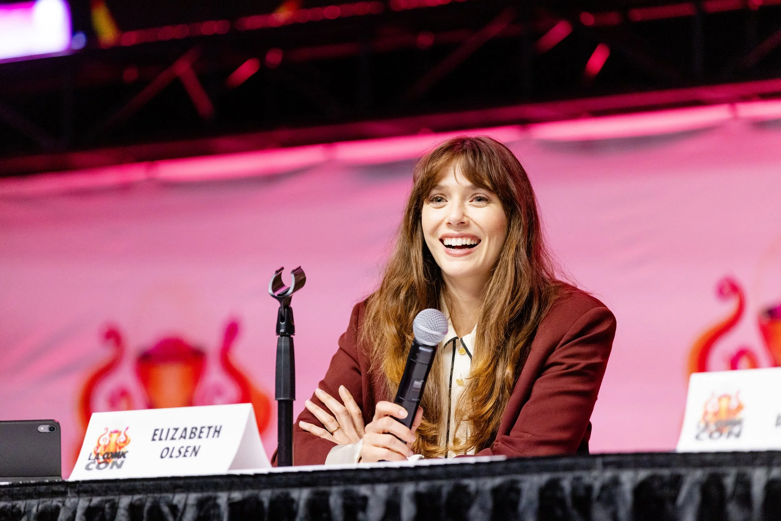 Person in a blue shirt speaking into a microphone at a comic convention, with a colorful graphic in the background.