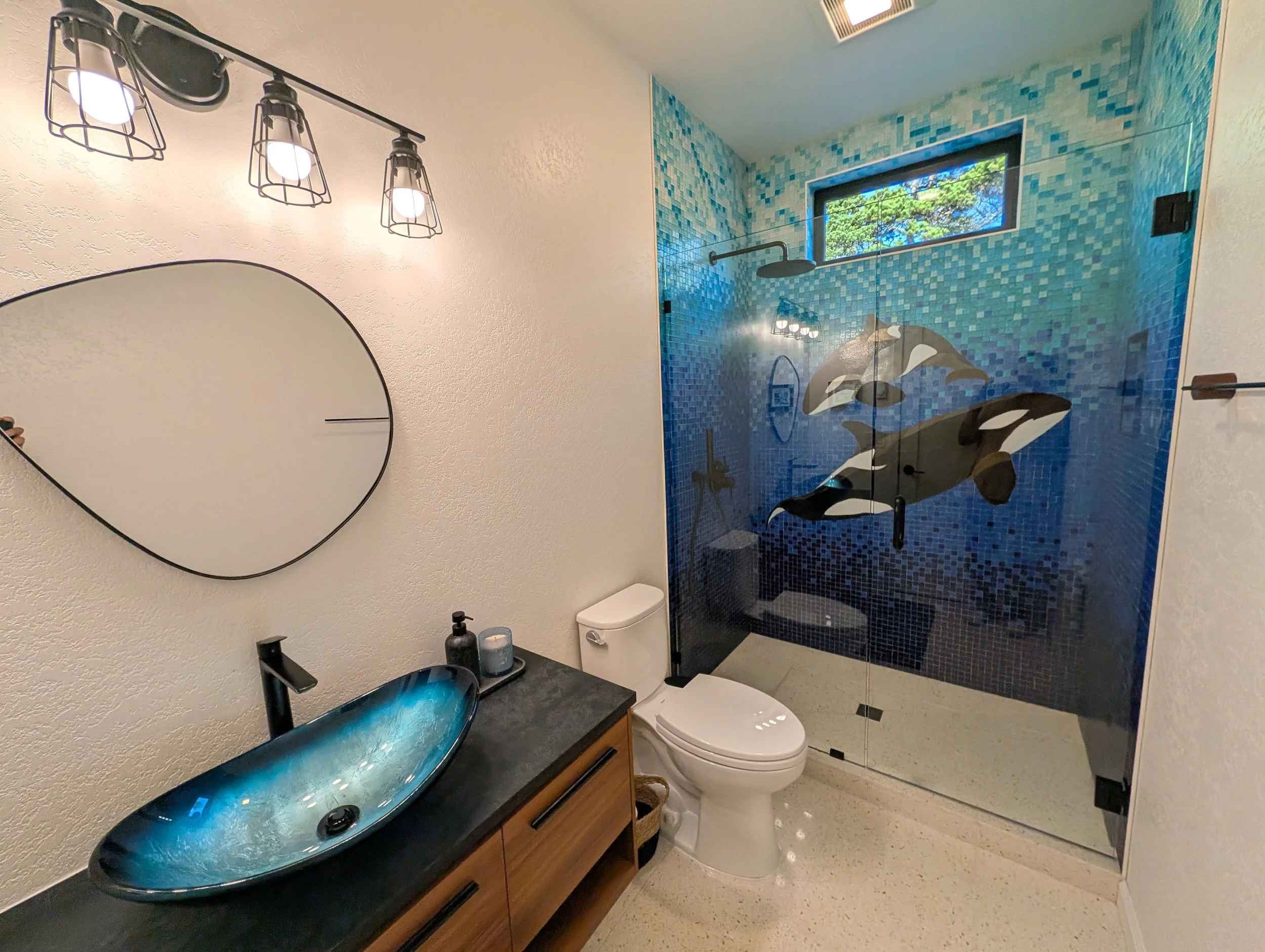Bathroom with a blue mosaic shower wall, glass shower door with orca whale design, window showing trees, modern black sink with wooden cabinet, toilet, and a mirror.