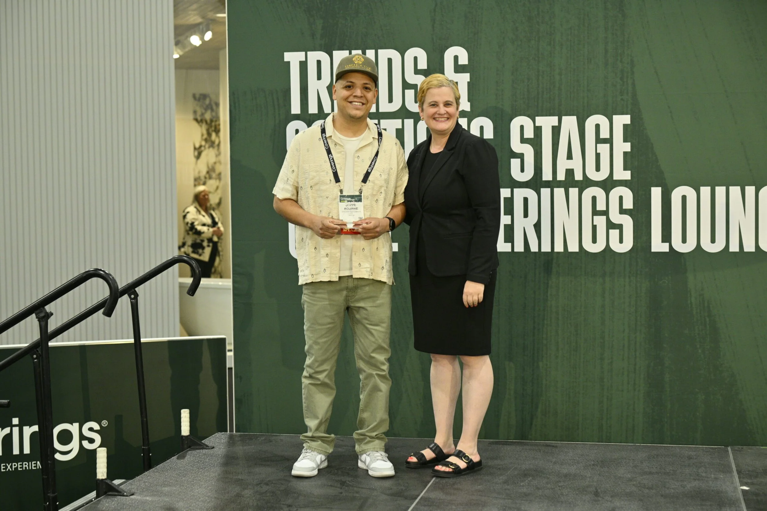 Two people standing on a stage at a conference, smiling at the camera. The person on the left is a young man wearing a beige patterned shirt, khaki pants, white sneakers, and a gray cap. The person on the right is a woman with short blonde hair, dressed in a black blazer, black skirt, and black sandals.