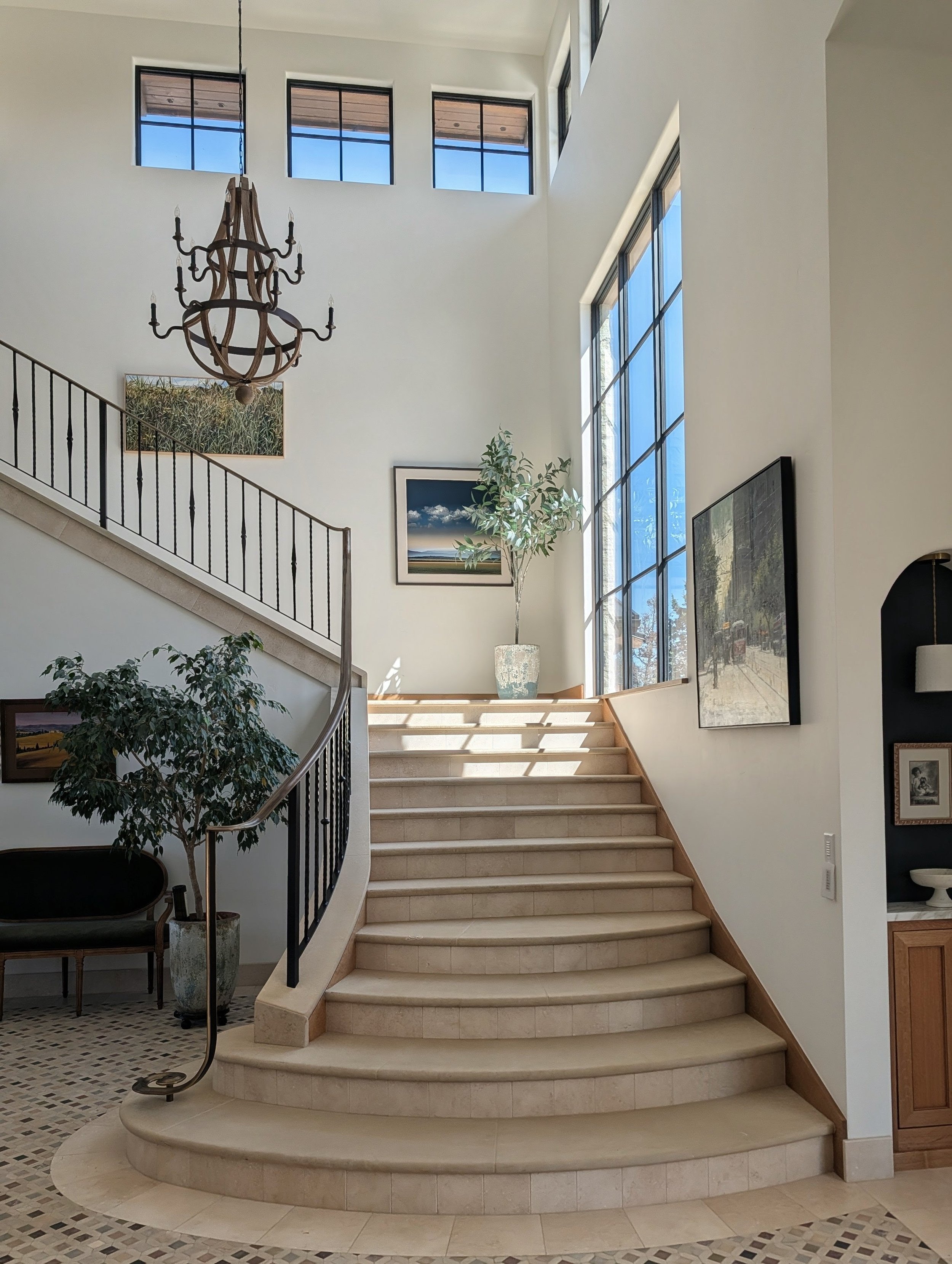 Interior of a modern building with a staircase, large windows, a chandelier, potted plants, and framed artwork on the walls.