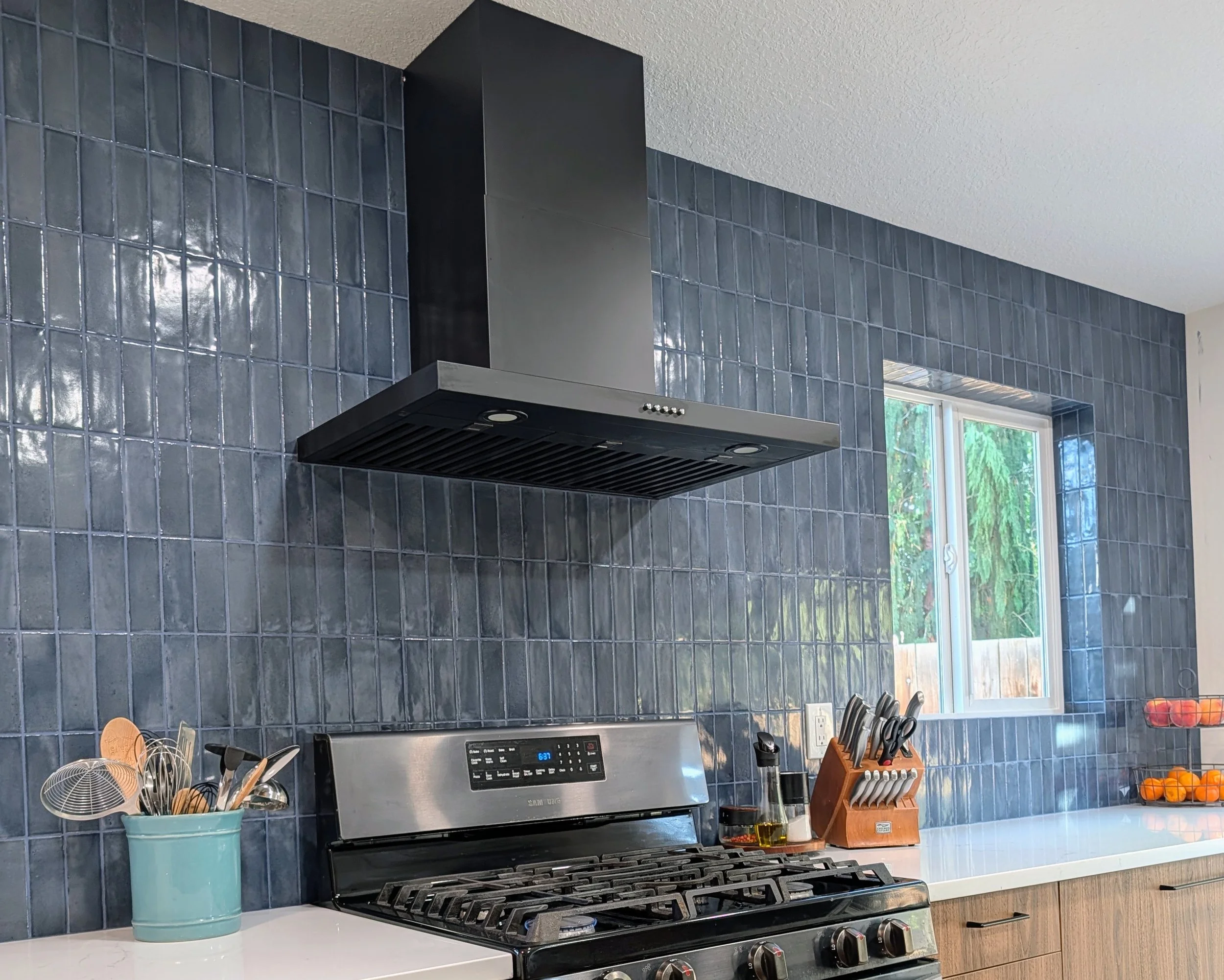 Kitchen with blue tiled backsplash, stainless steel range hood above stove, and window showing greenery outside. Counter has utensils in a light blue container, a knife block, and a bowl of oranges.