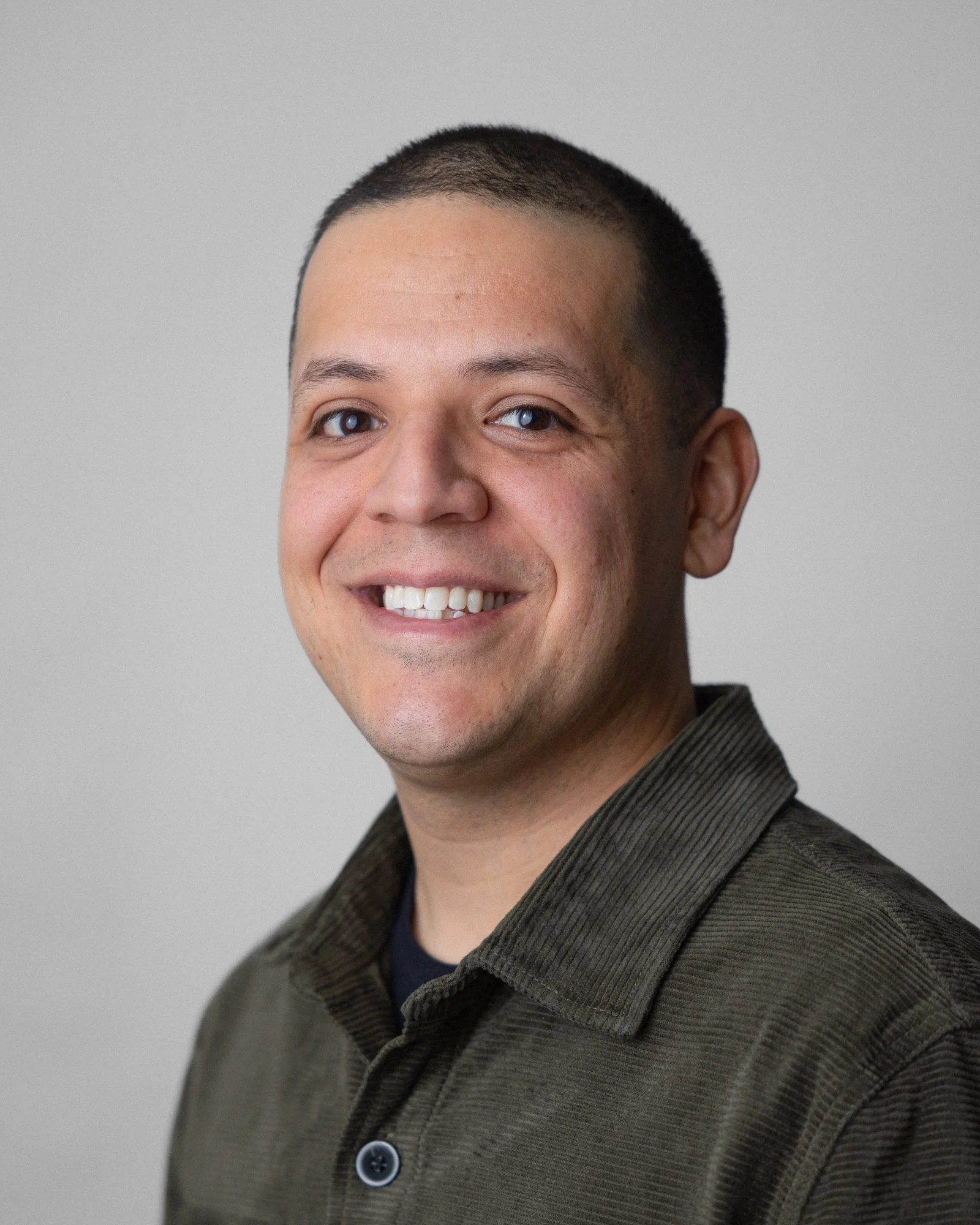 Close-up of a man with short dark hair and brown eyes, smiling, wearing a dark collared shirt and blazer, against a plain light gray background.