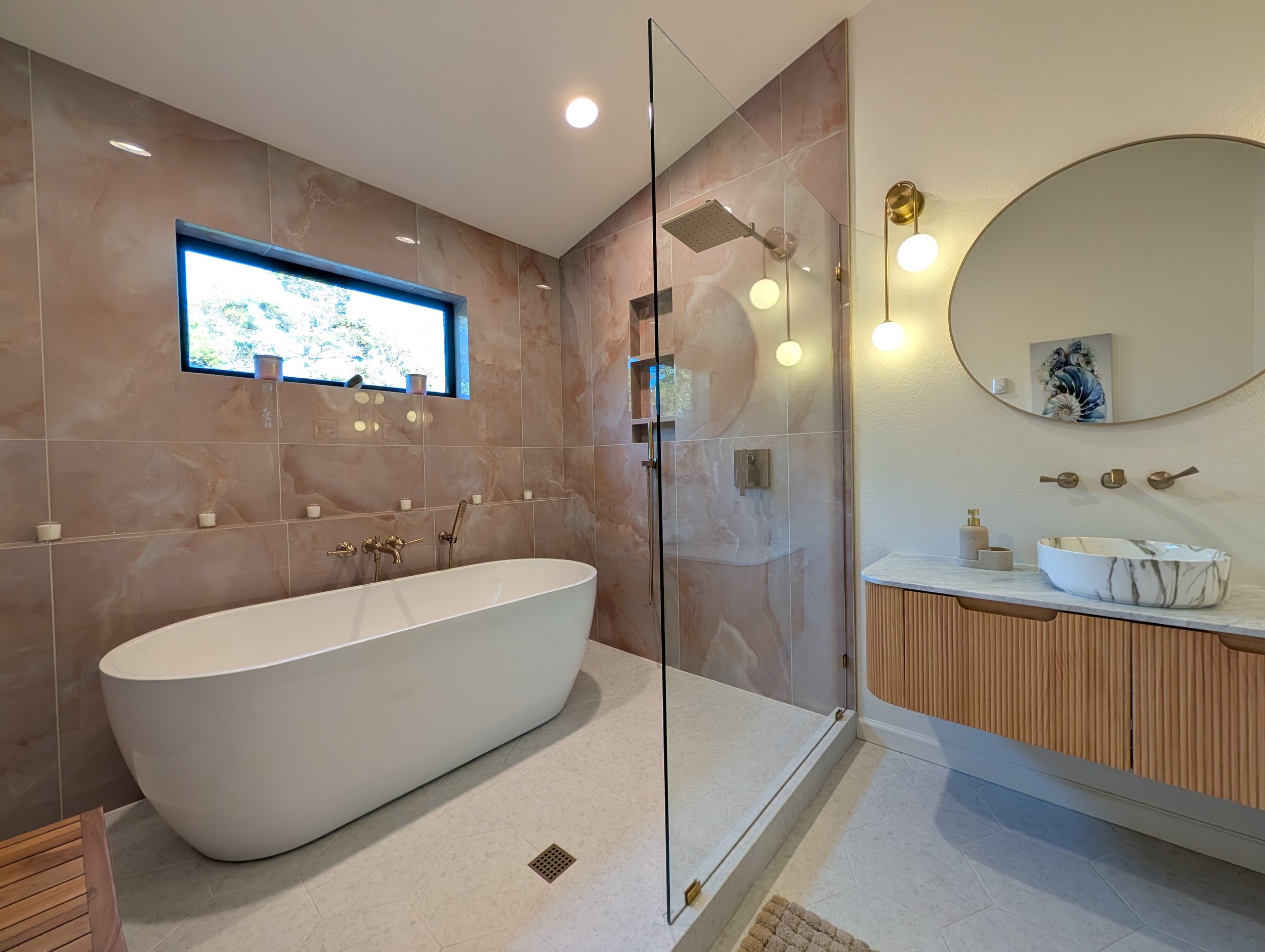 Modern bathroom with a white freestanding bathtub, a shower with glass enclosure, marble wall tiles, a wall-mounted round mirror, and a vanity with a marble sink and wooden cabinet, illuminated by spherical pendant lights.