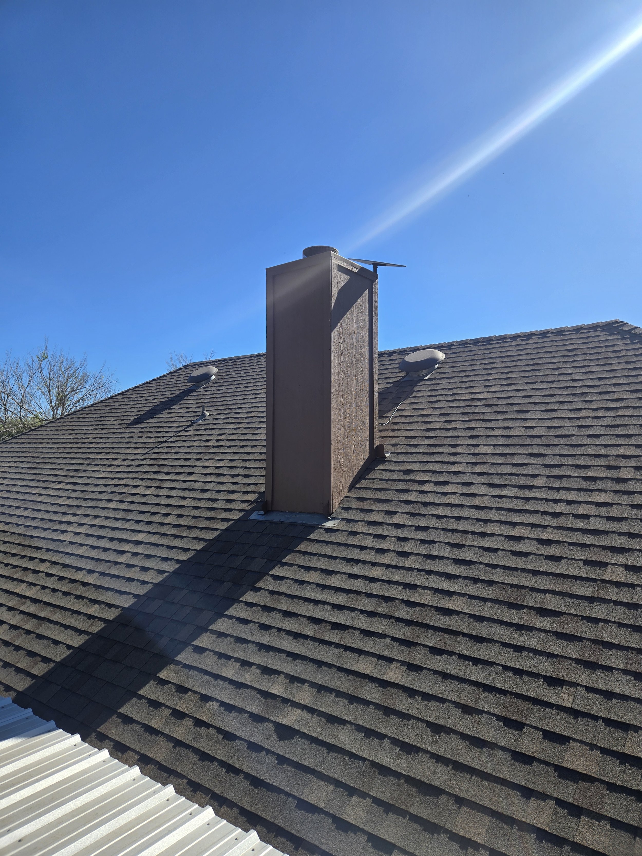Roof with brown shingles, two circular roof vents, a chimney with a small weather vane, a blue sky, and some trees in the background.