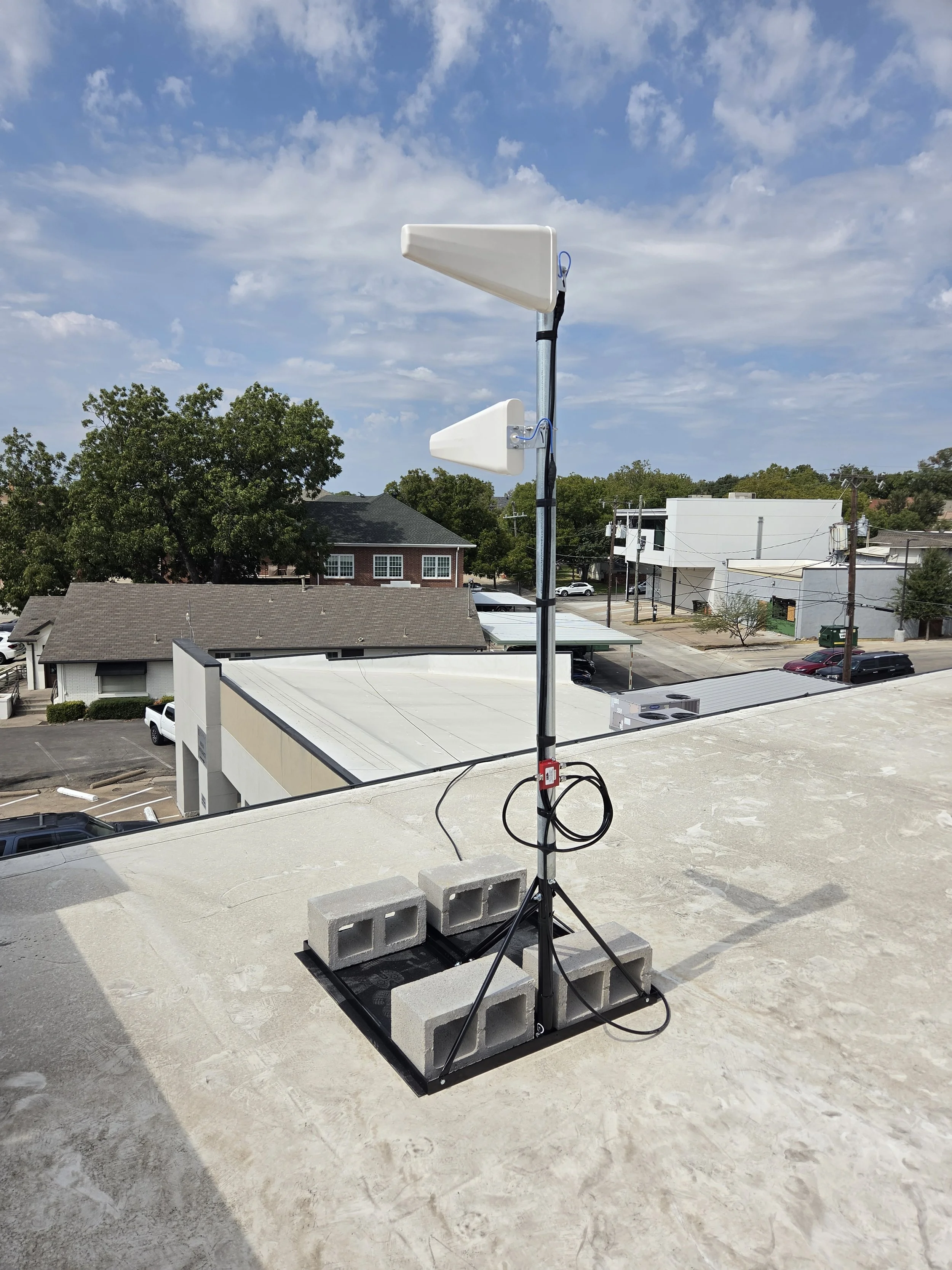 A rooftop with a metal pole supporting two large white antennas, surrounded by concrete blocks and electrical wiring, overlooking a neighborhood with houses, trees, and parked cars under a partly cloudy sky.