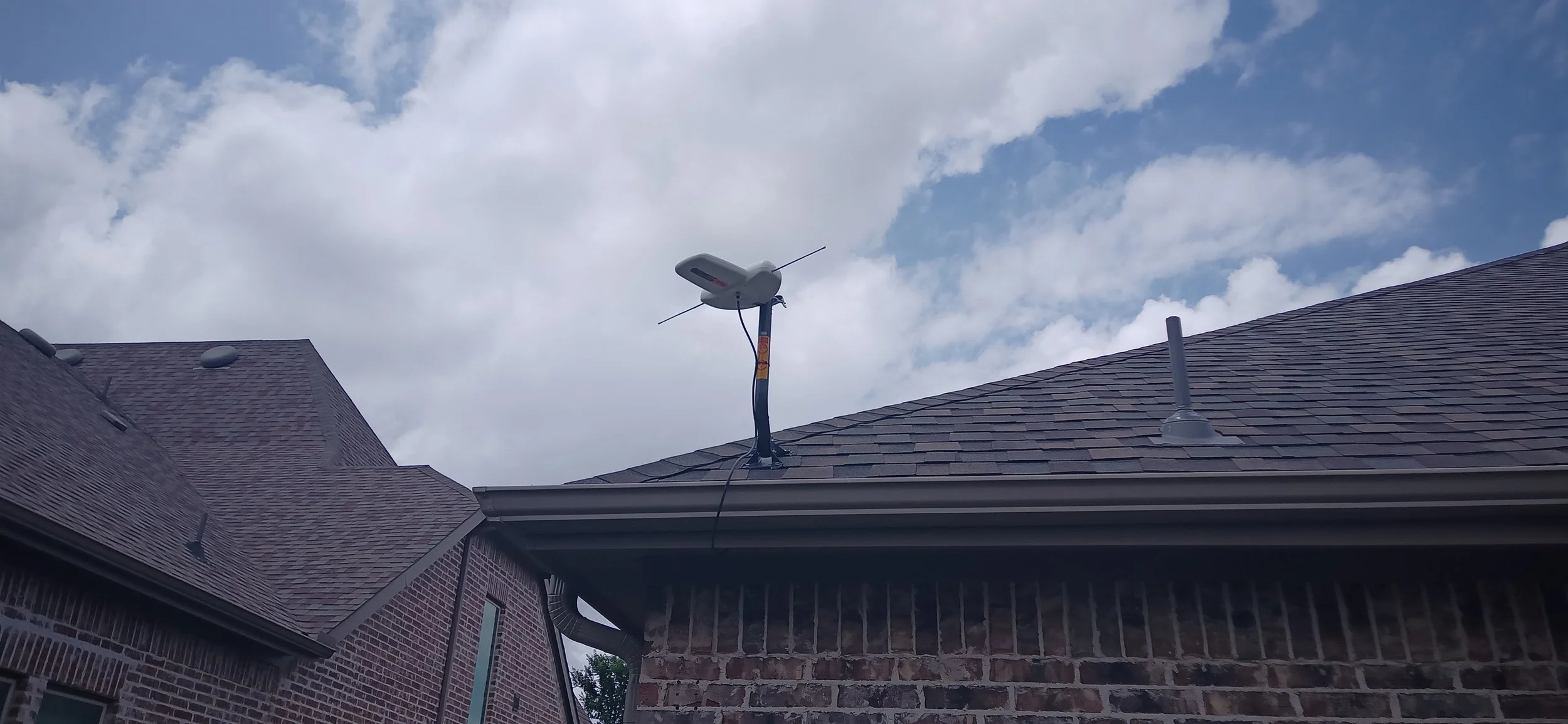 View of a house roof with an antenna and weather sensor mounted on it, under a partly cloudy sky.