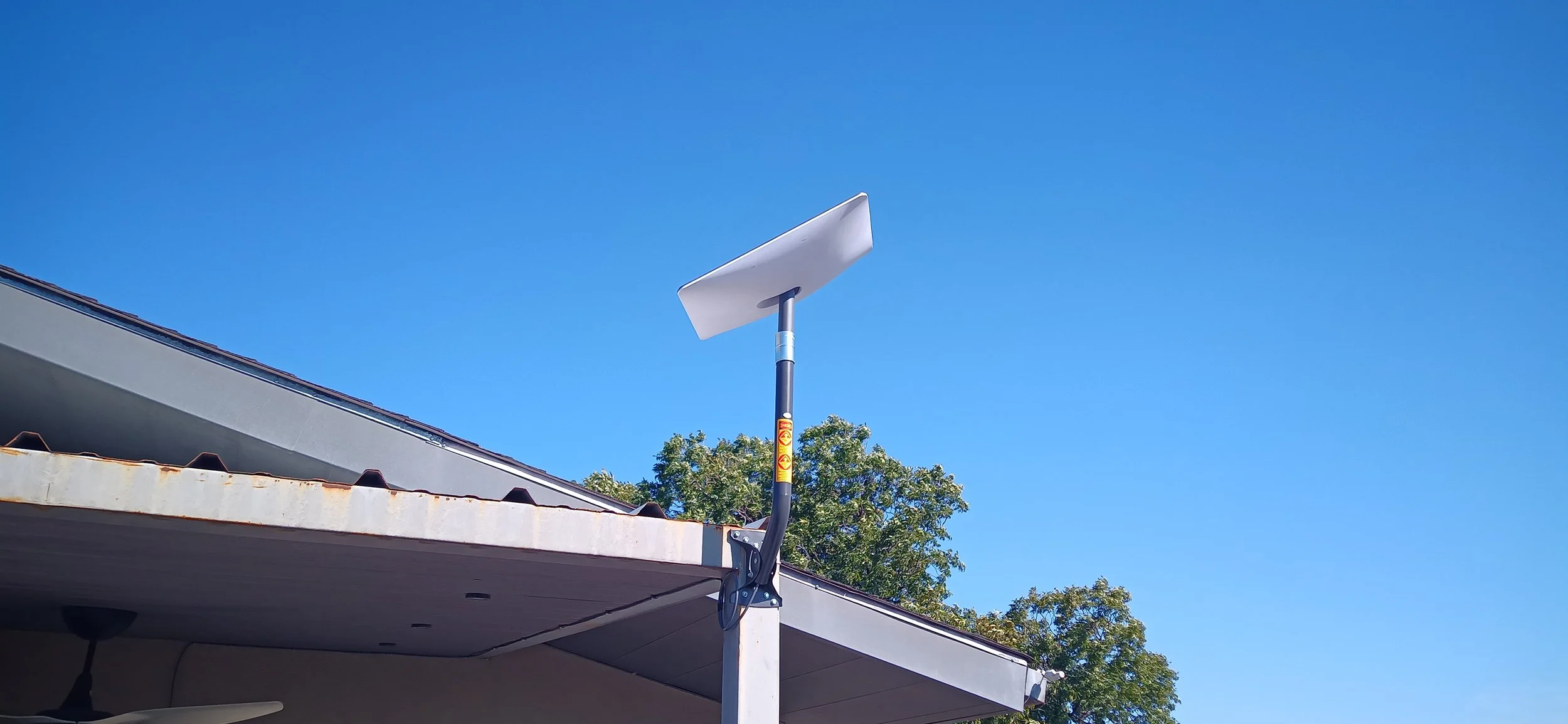 Outdoor scene with a blue sky, part of a house's roof, a solar panel on a pole, and trees in the background.