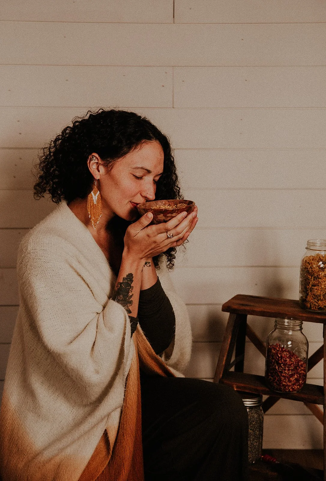 Woman with curly dark hair and earrings holds a bowl close to her nose, appearing to smell its contents, sitting indoors against a light wooden wall with jars of herbs on a small wooden shelf beside her.