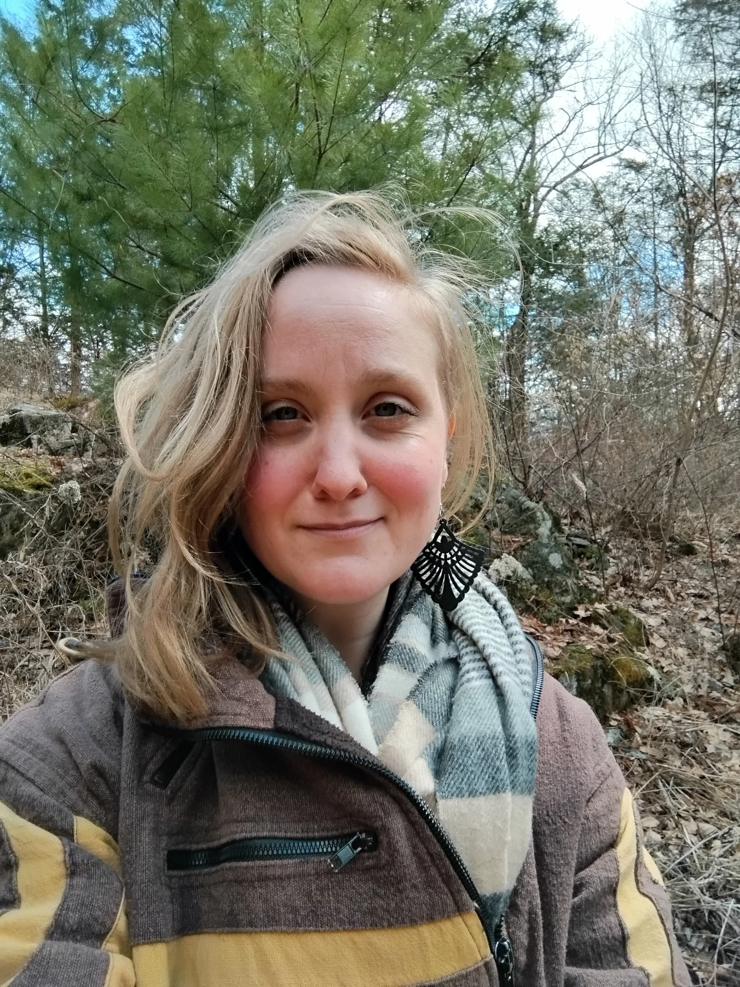A woman with blonde hair smiling outdoors in a wooded area during daytime, wearing a brown and yellow jacket, a plaid scarf, and a black earring.