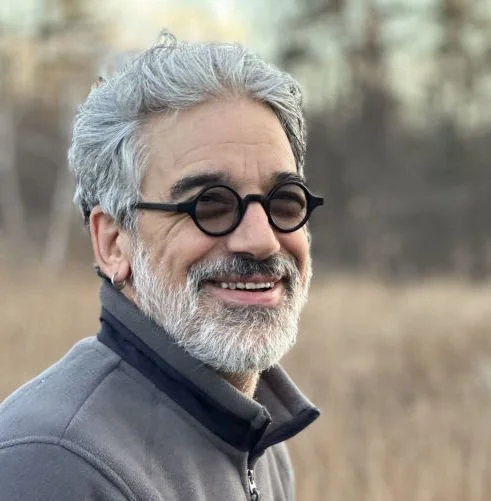 A smiling middle-aged man with gray hair, a beard, wearing black glasses and a gray jacket outdoors.