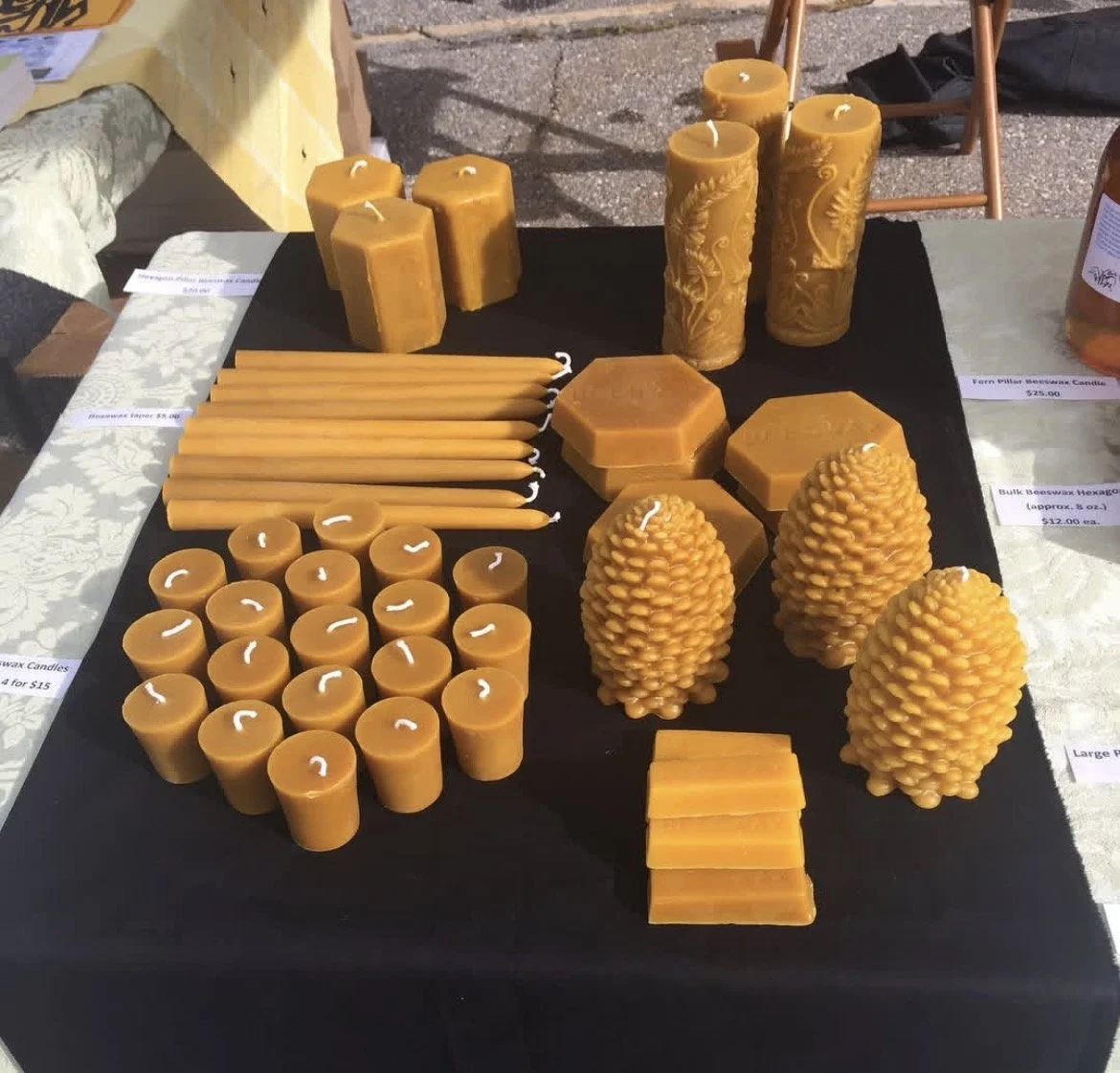 A display of yellow beeswax candles in various shapes and sizes, including hexagons, cylinders, cones, and textured pine cone-like designs, on a black table at a market.