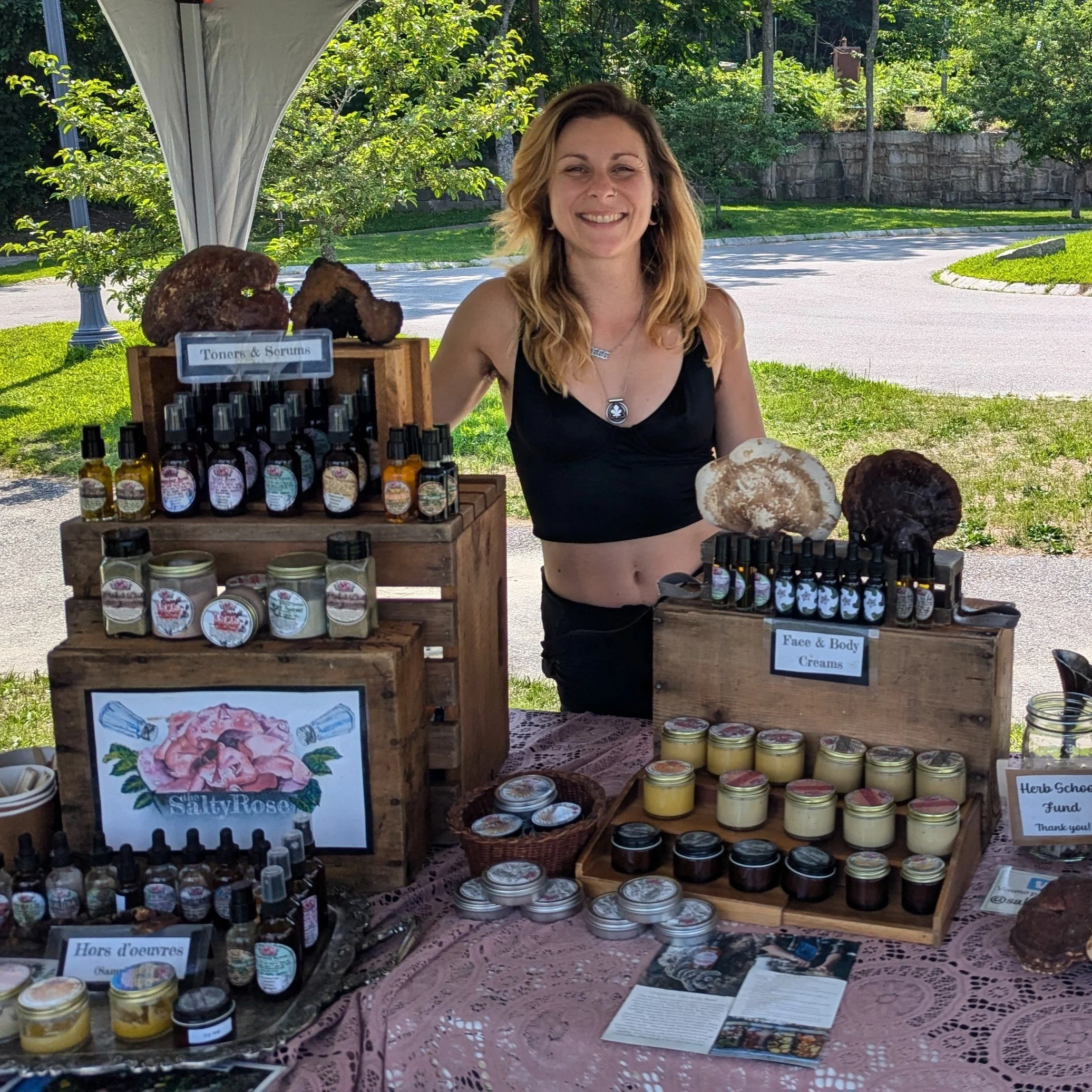 A woman standing behind a market stall booth with handmade herbal products, including creams, oils, and salves, set outdoors on a sunny day. The booth displays jars and bottles on wooden crates with signs indicating various product categories, and there are large decorative mushrooms on the display.