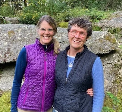 Two women smiling outdoors in front of rocks and greenery, one wearing a purple quilted vest and the other in a navy vest.
