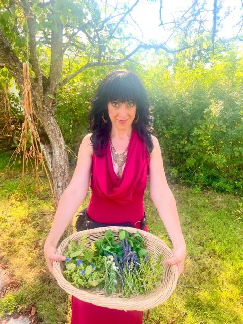 Woman wearing a red dress holds a basket of fresh herbs and greens outdoors in a sunny garden with trees and greenery in the background.