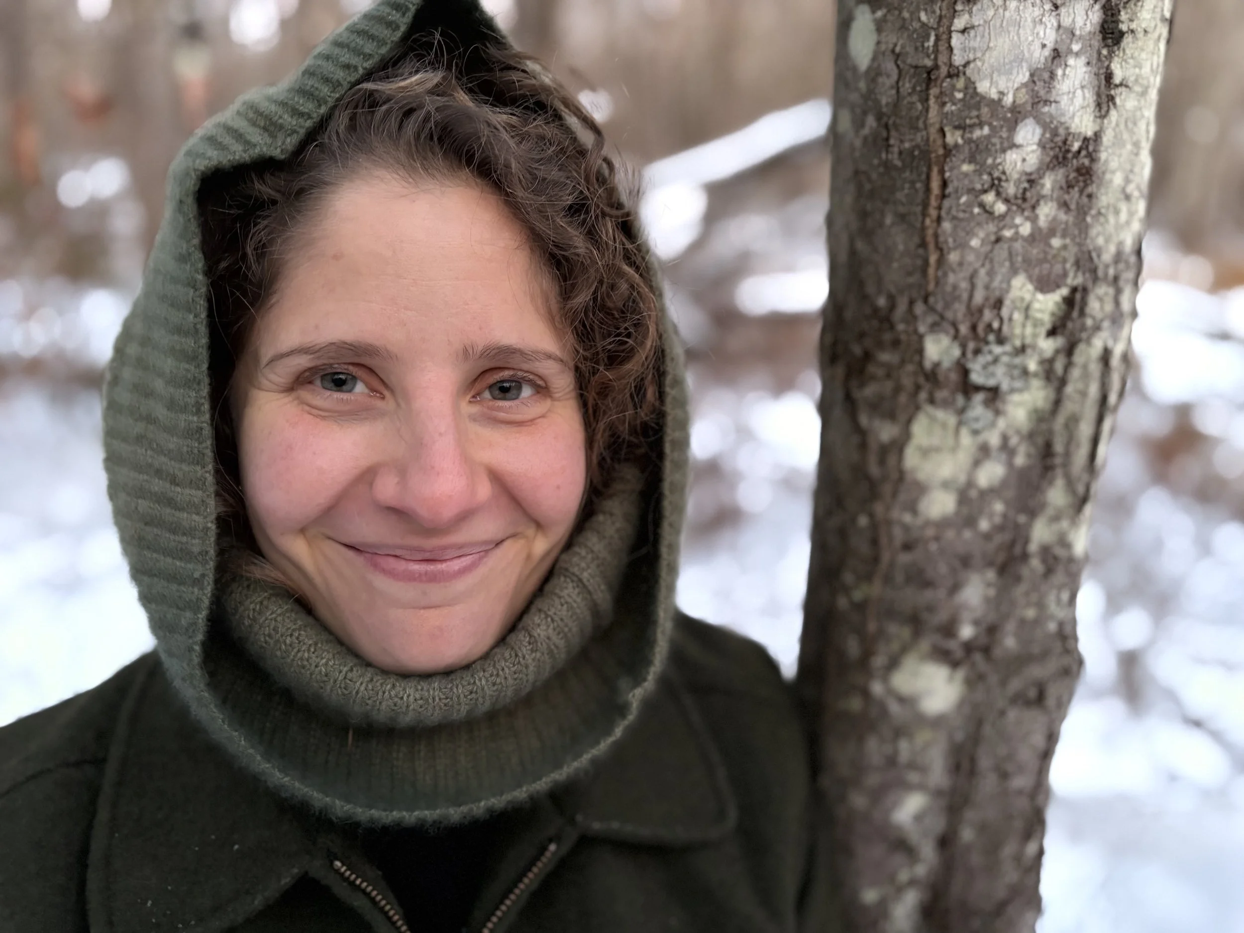 A woman smiling in a winter forest, wearing a hooded coat and standing next to a tree with snow on the ground.