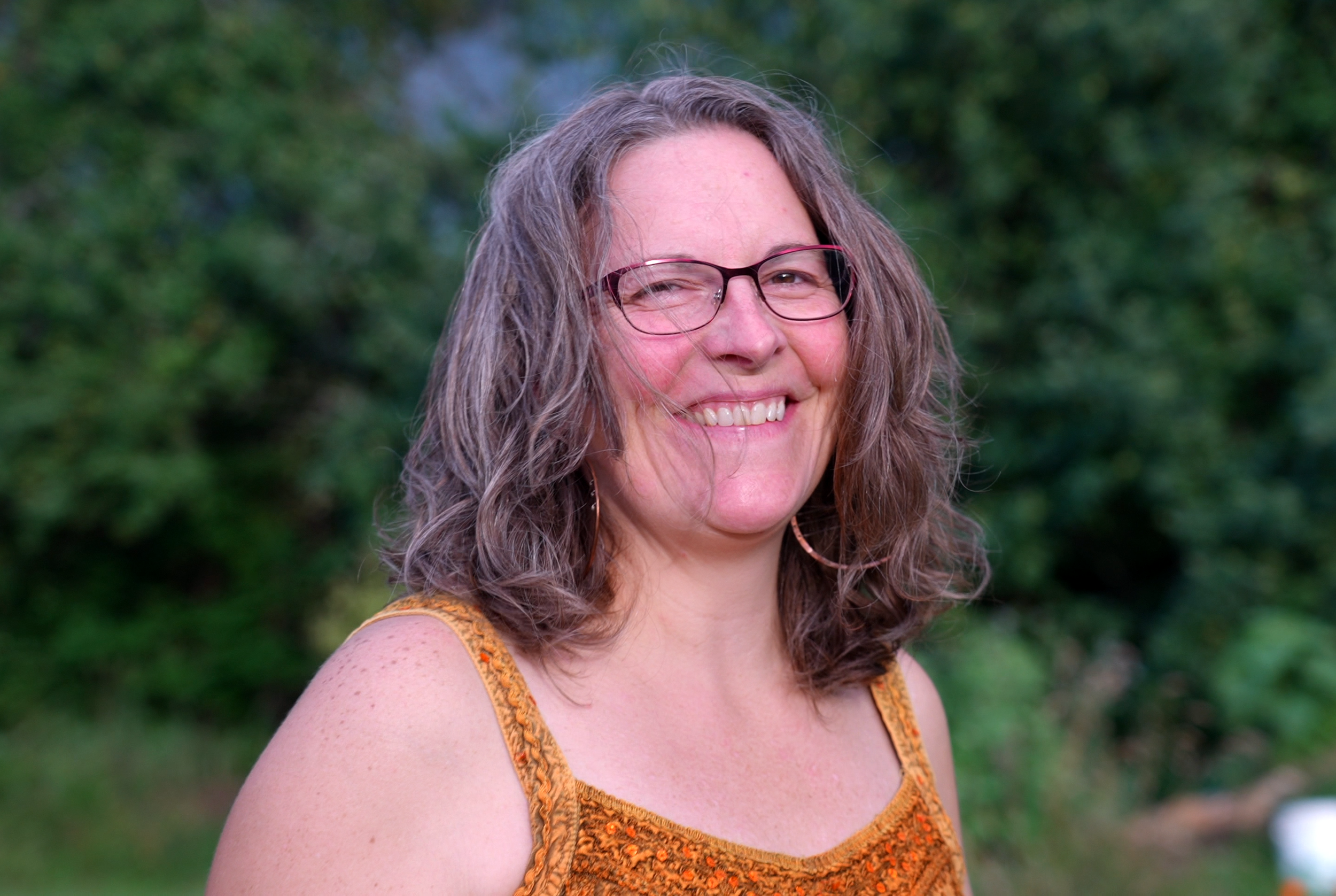 A woman with curly hair, wearing glasses and a sleeveless orange top, smiling outdoors with a green, wooded background.