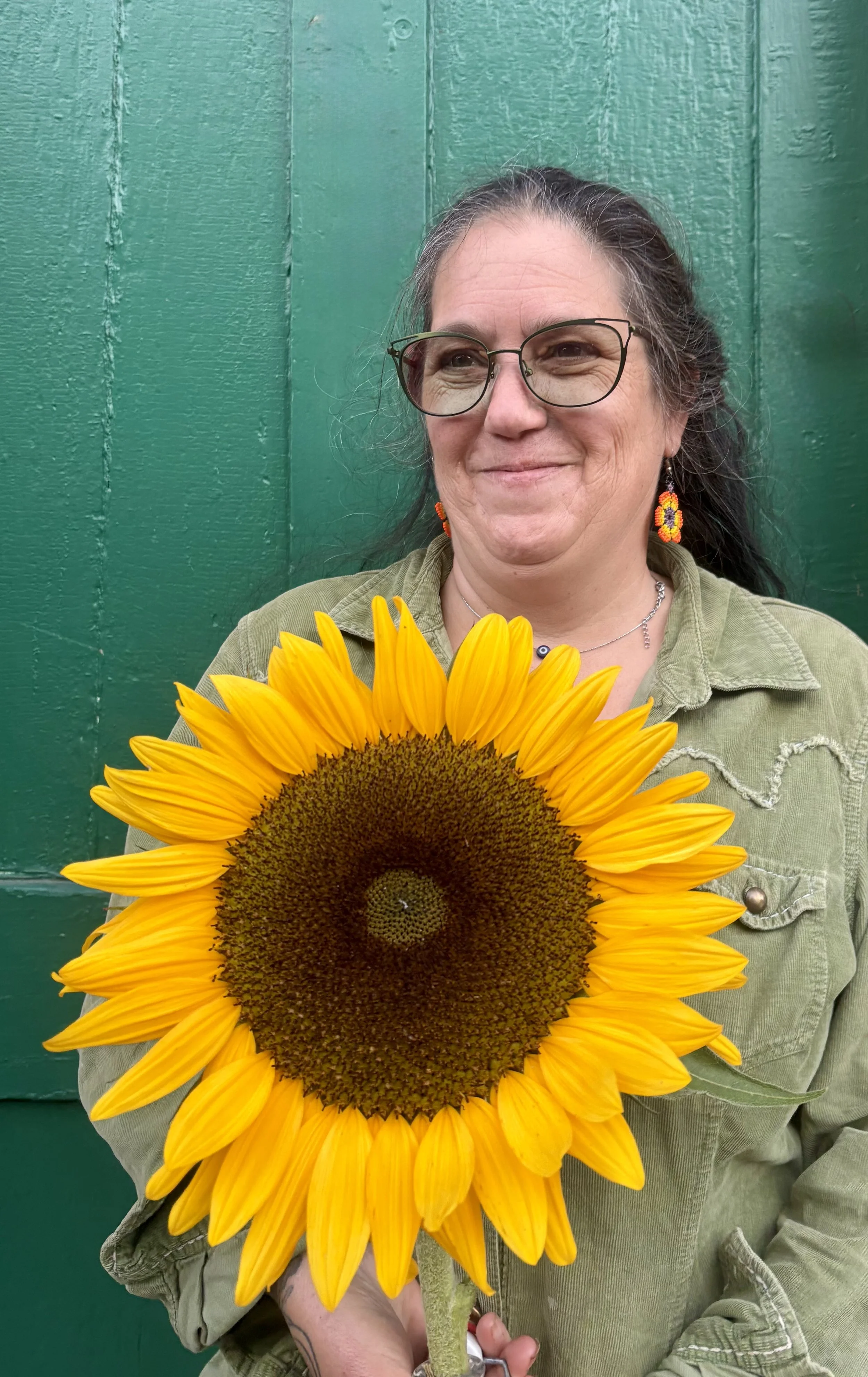 A woman with glasses, long dark hair, and earrings, standing in front of a green wooden background, holding a large sunflower.