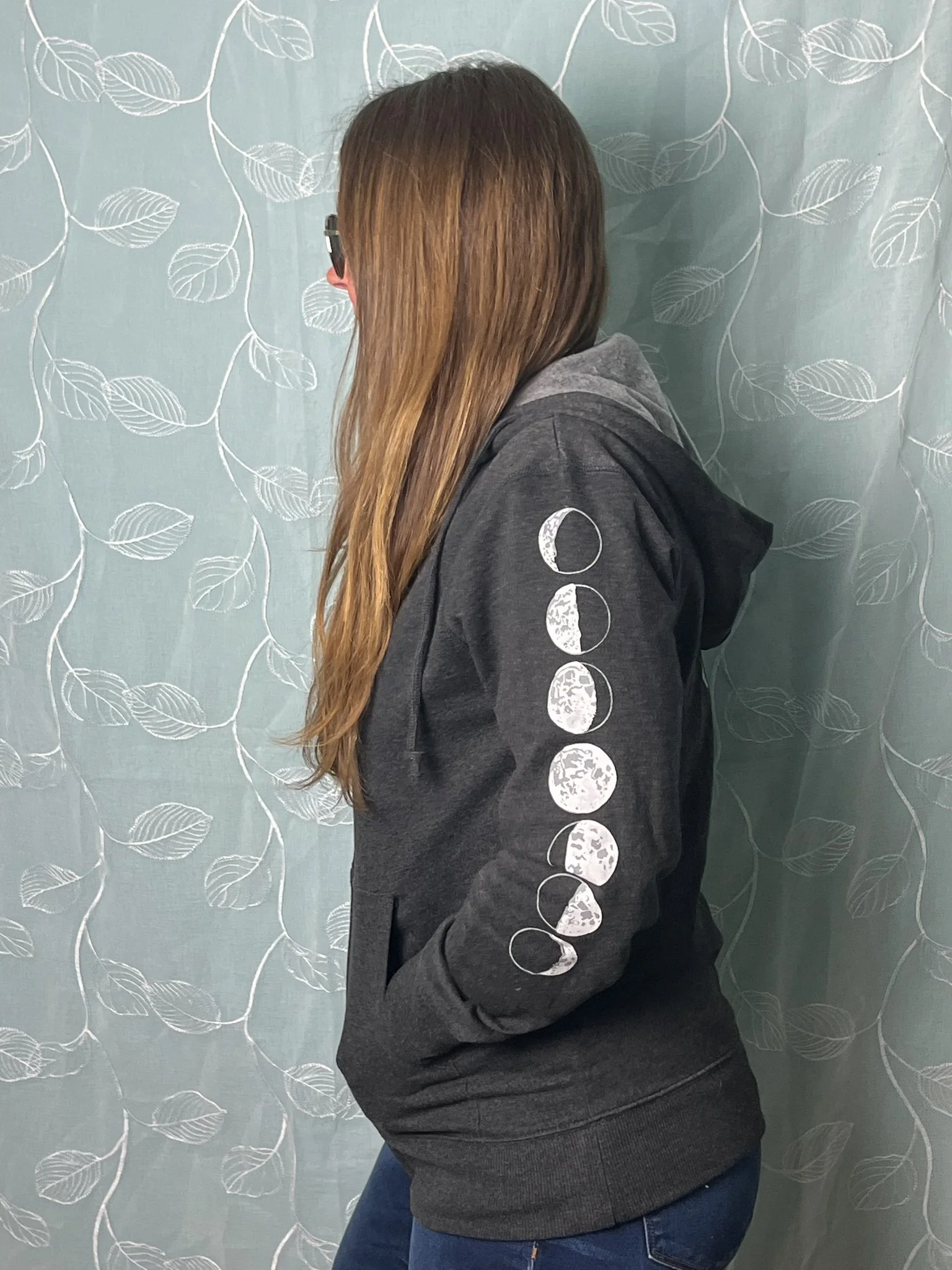 A woman with long hair wearing a black hoodie with moon phases printed on the sleeve, standing against a light blue wall with white leaf patterns.