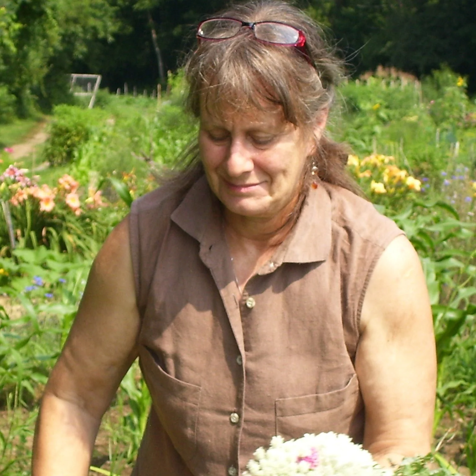 A woman with brown hair and glasses on her head, wearing a sleeveless brown shirt, stands in a garden with colorful flowers, holding a bouquet of white flowers and smiling.