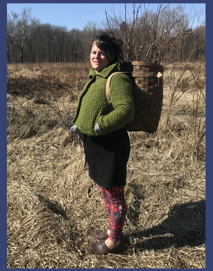 Woman outdoors in a field with dry grass and leafless trees, wearing a green jacket, black skirt, colorful leggings, and brown shoes, carrying a large woven basket on her back filled with sticks or branches.