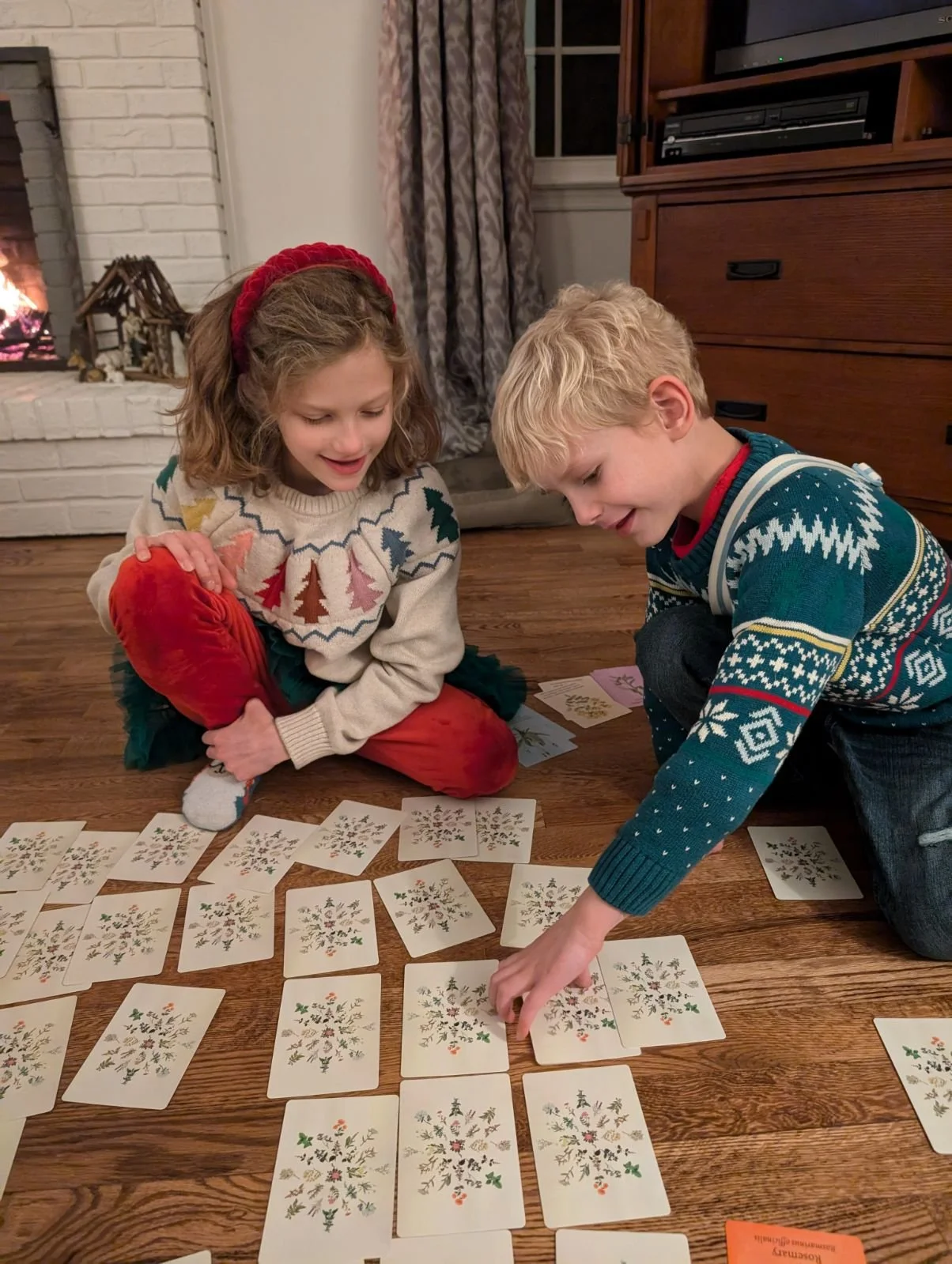 Two children playing a holiday-themed card game on a wooden floor near a fireplace. The girl wears a sweater with a tree pattern and a red headband, while the boy wears a Christmas sweater with a snowflake pattern.