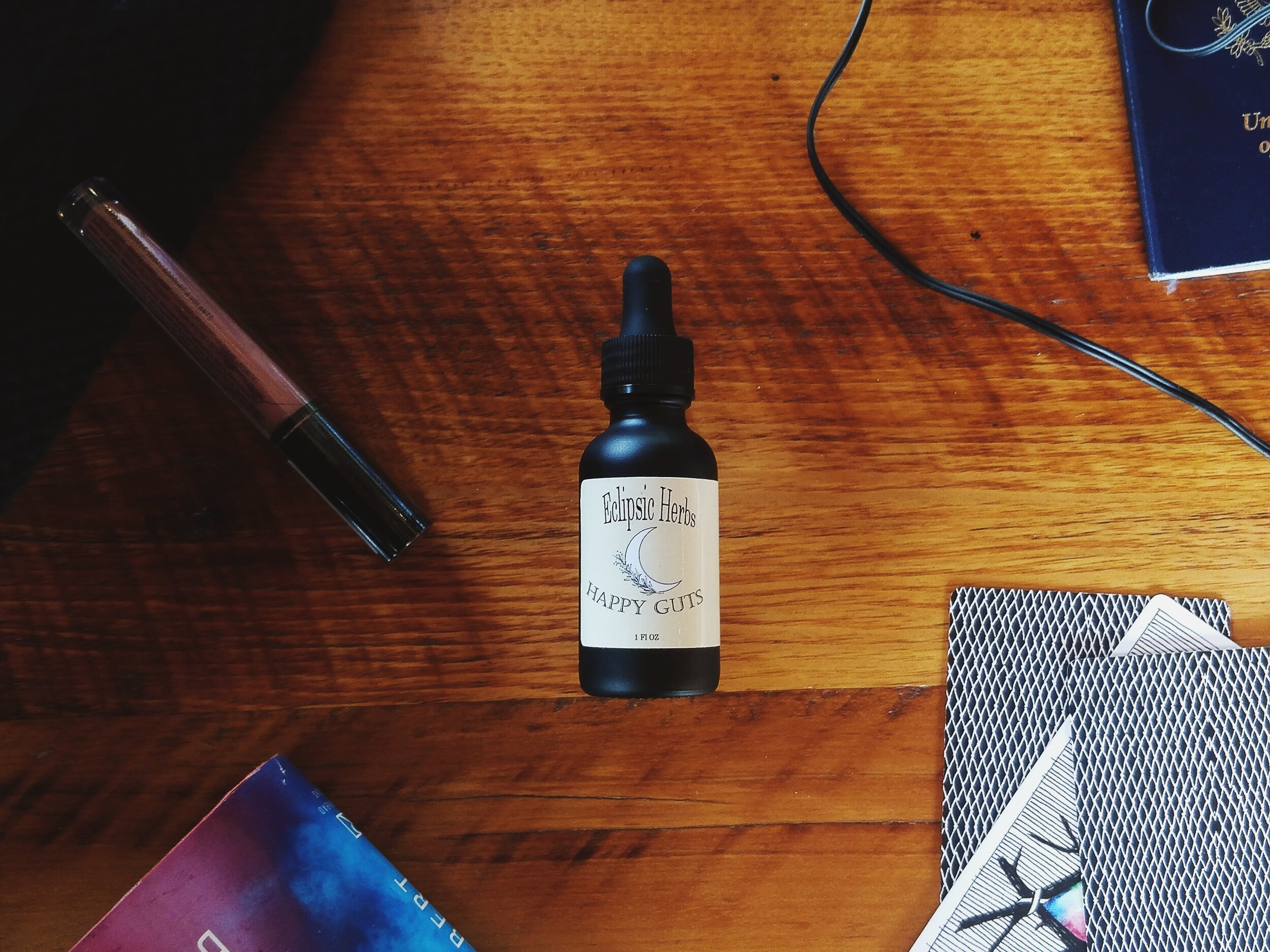 A small black bottle labeled 'Ecliptic Herbs Happy Guts' placed on a wooden surface with various objects around it, including a notebook, playing cards, a pen, and a wire.