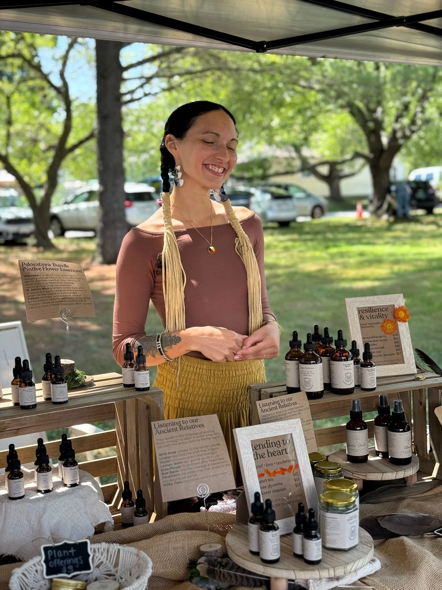 Woman standing behind a display of essential oils and herbal products at an outdoor market, under a canopy with trees and cars in the background.