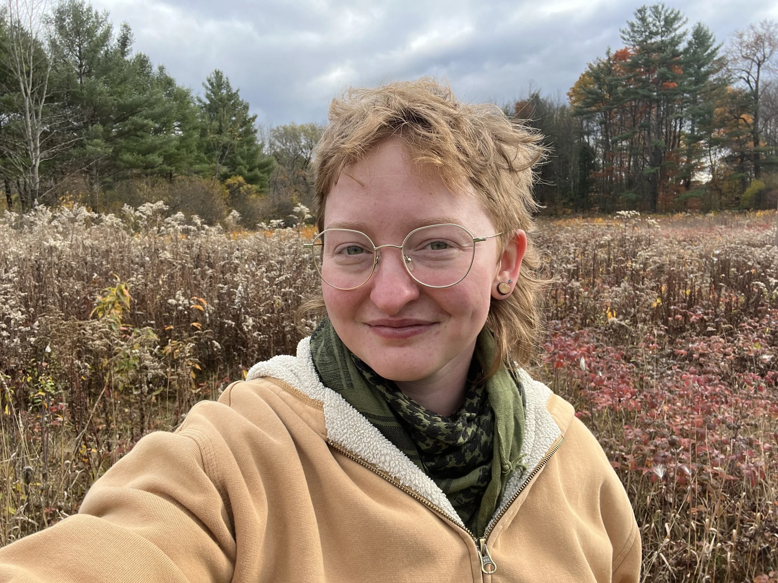 A person with red hair, glasses, and earrings taking a selfie outdoors in a field during fall with trees showing autumn colors in the background.