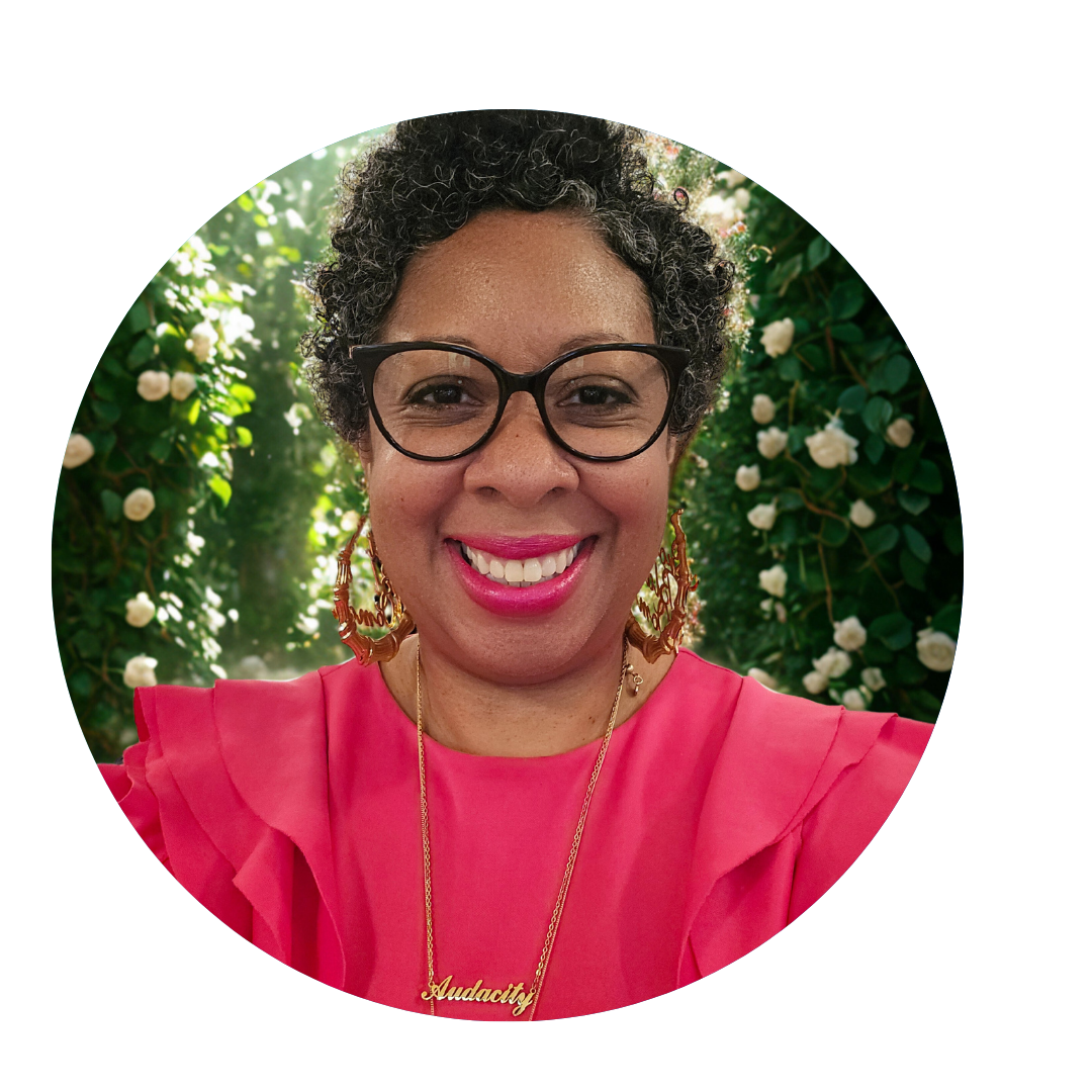 A smiling woman with short curly hair, wearing large glasses, gold earrings, and a pink top, standing in front of a background with greenery and white flowers.