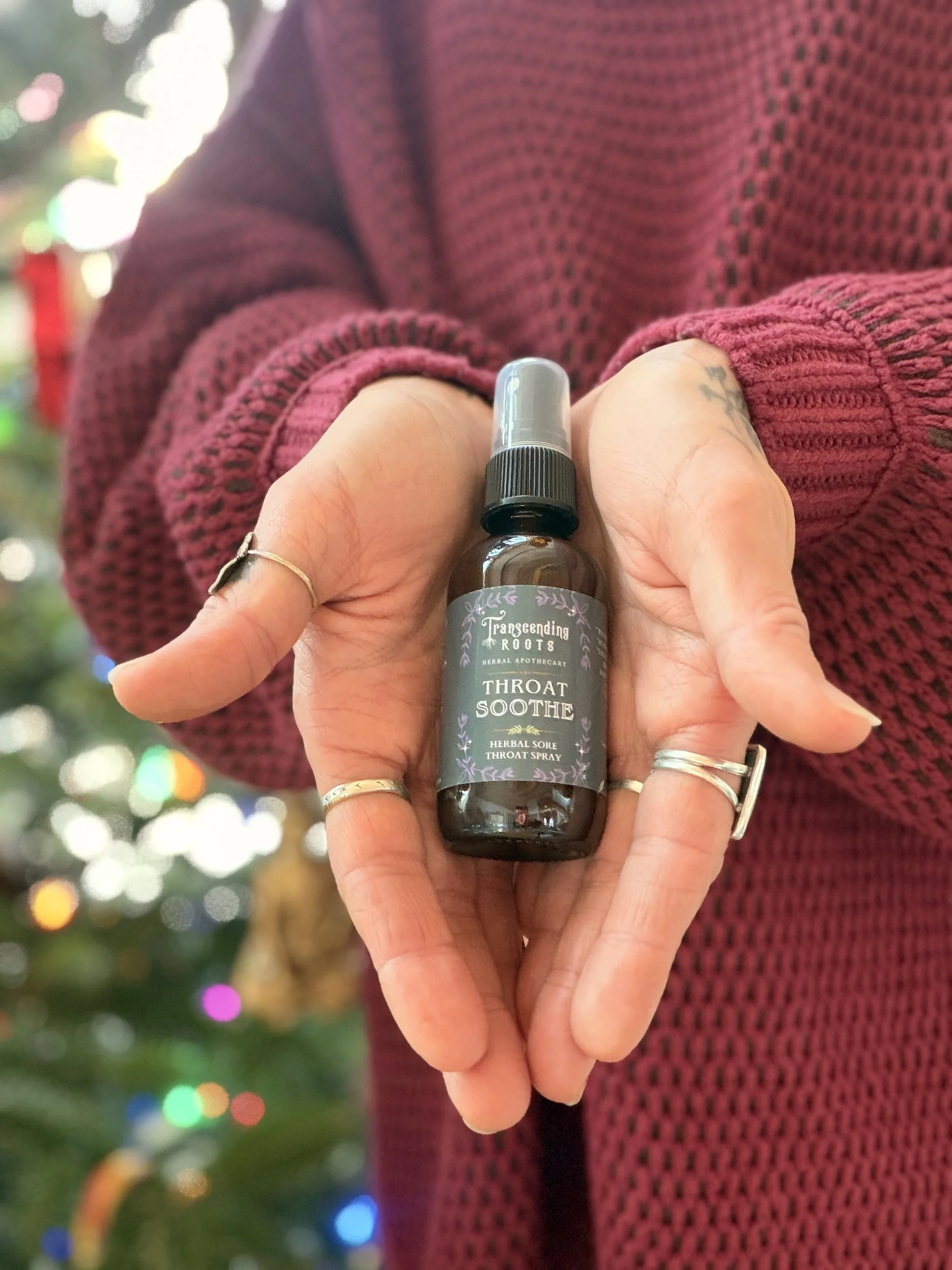 A person holding a small bottle of herbal throat spray labeled 'Transcending Roots Throat Soothe' in front of a decorated Christmas tree.