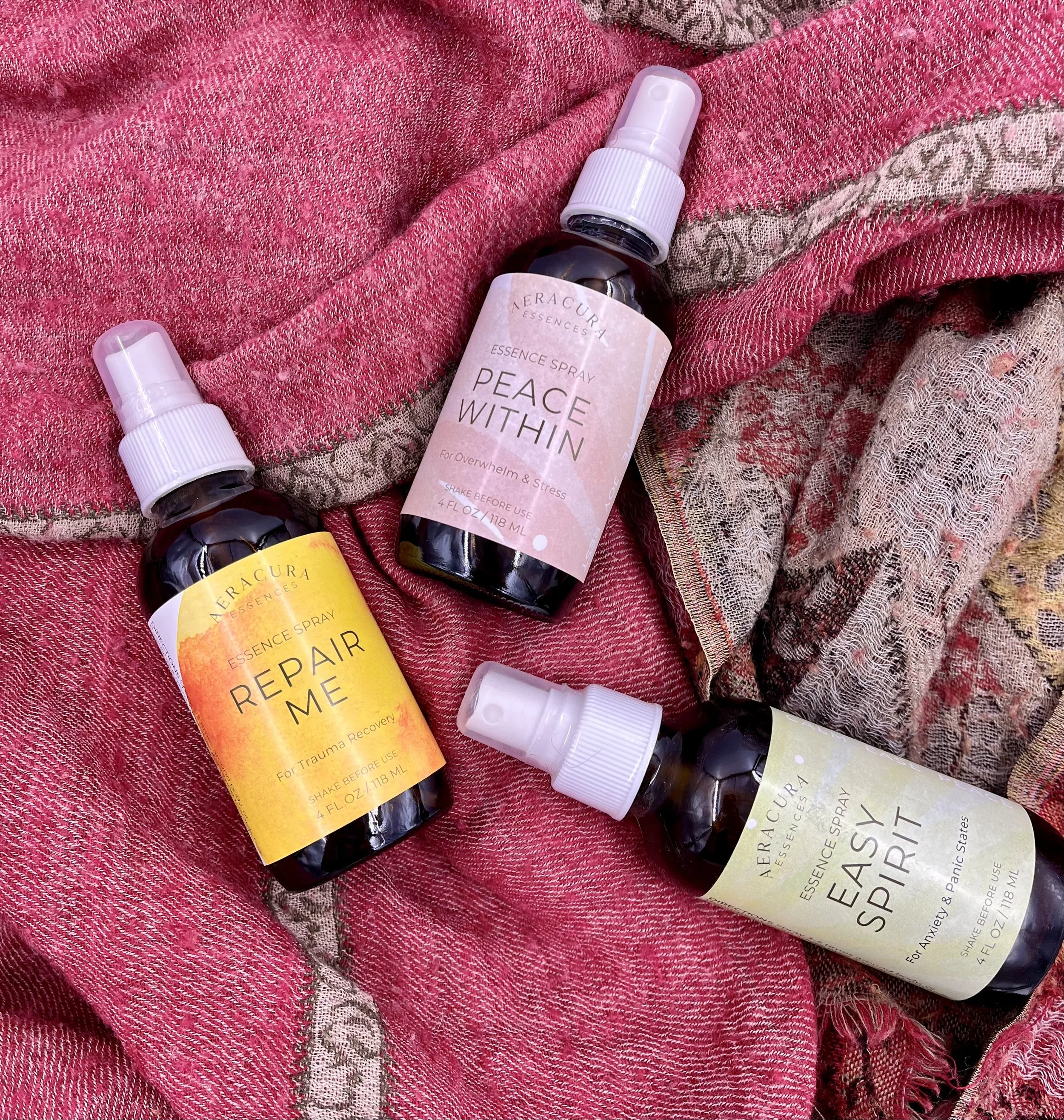 Three spray bottles with labels reading Repair Me, Peace Within, and Easy Spirit on a pink and beige textured fabric.