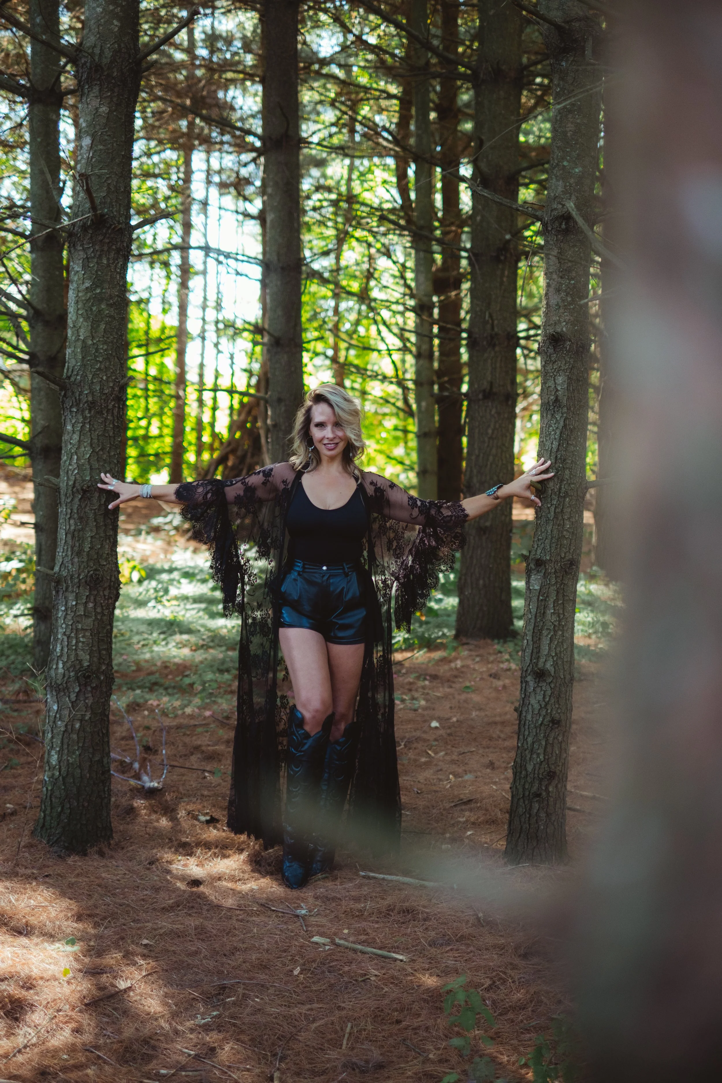 A woman in a black outfit and high boots standing in a forest with her arms outstretched, wearing a sheer black lace robe.
