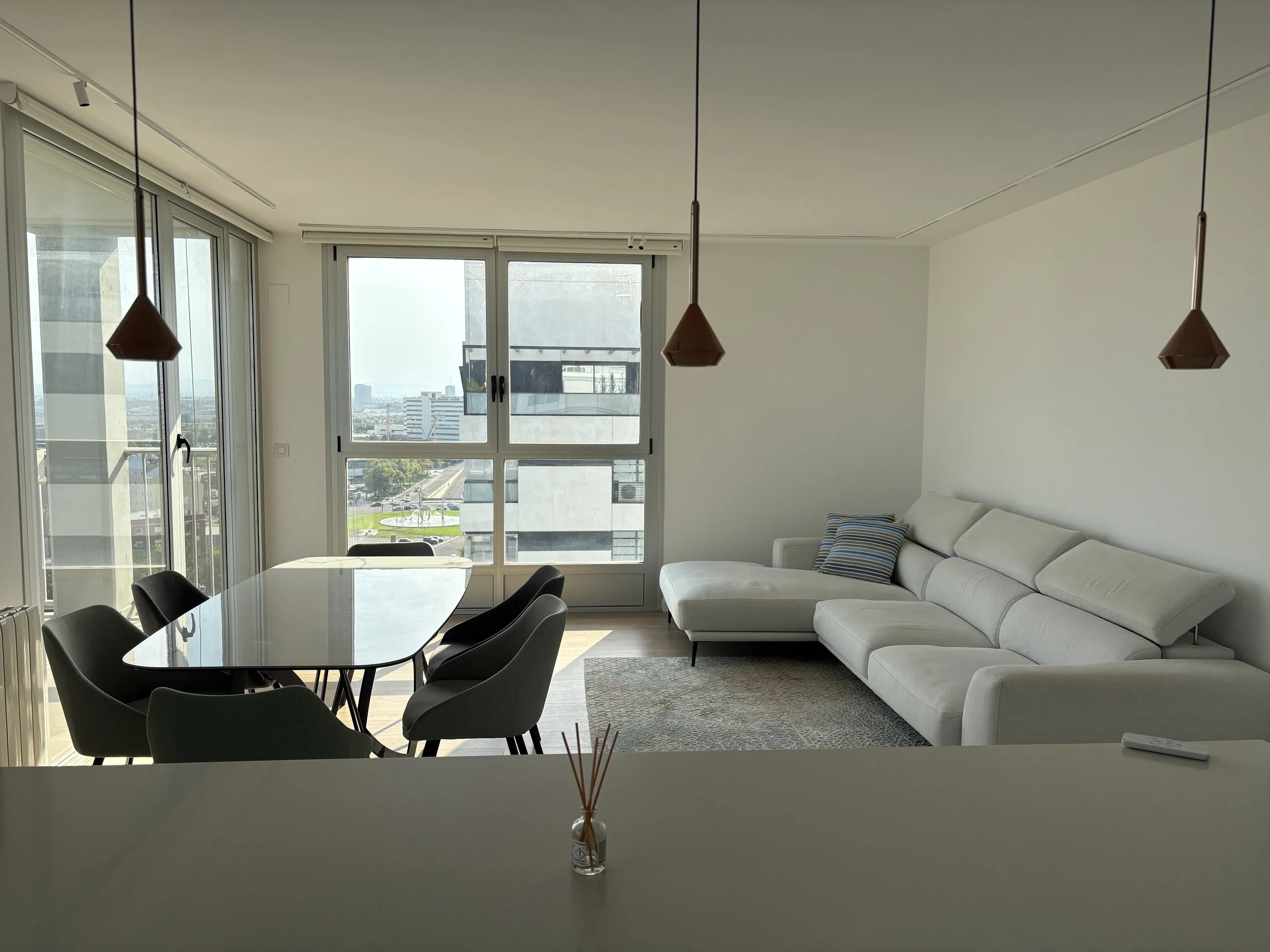 Sala moderna con sofá blanco, mesa de comedor ovalada con seis sillas negras, ventanas grandes con vista a la ciudad, lámparas colgantes de color marrón, alfombra gris y acento de cojín con rayas