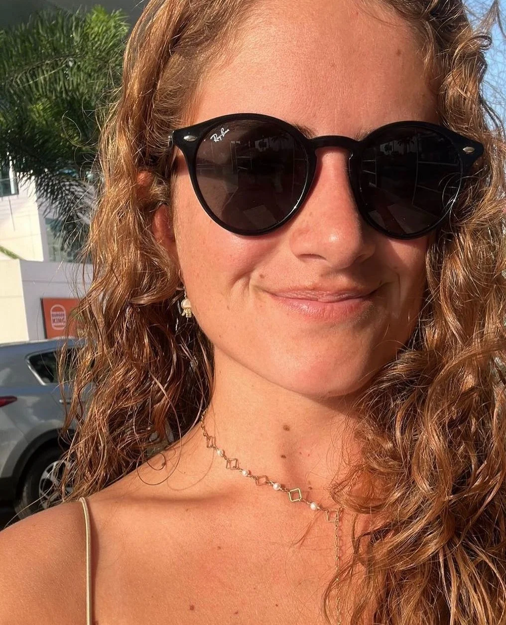 Mujer con cabello rizado, usando gafas de sol negras Ray-Ban, sonriente, con collares y ropa de tirantes, en un día soleado. En el fondo hay un auto y plantas tropicales.
