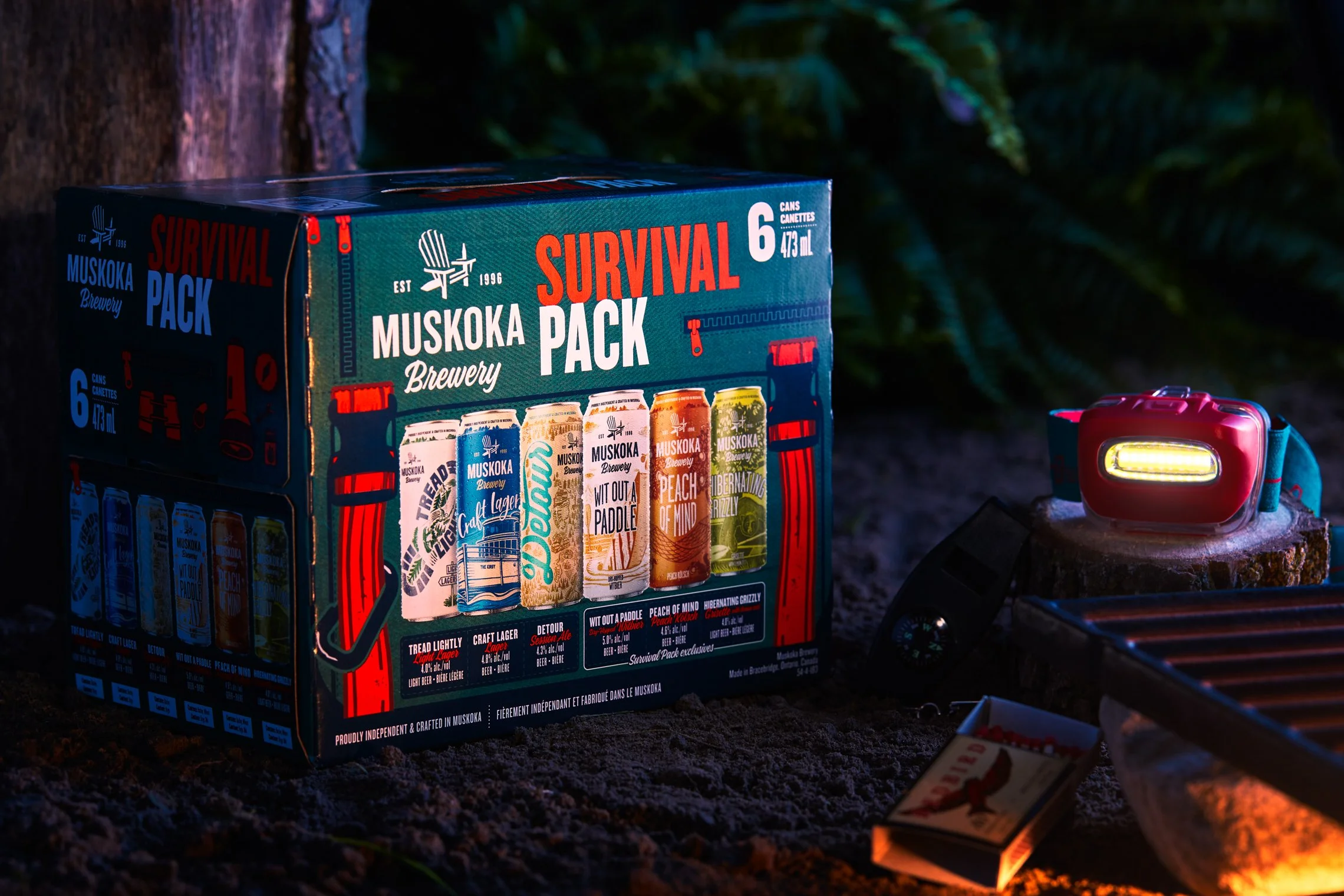 Muskoka Brewery Survival Pack box containing six cans of craft beer in a camping scene with a headlamp and fire pit.