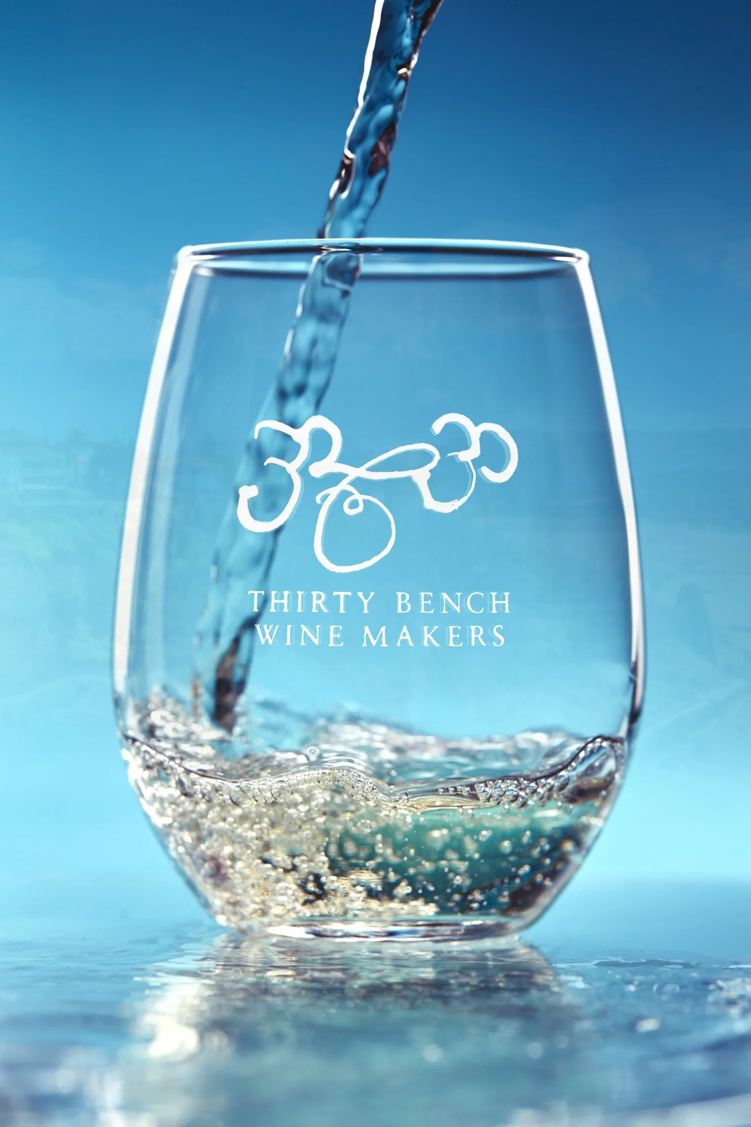 A glass of water being filled from above, with a logo and the text "Thirty Bench Wine Makers" on it, set against a blue background.