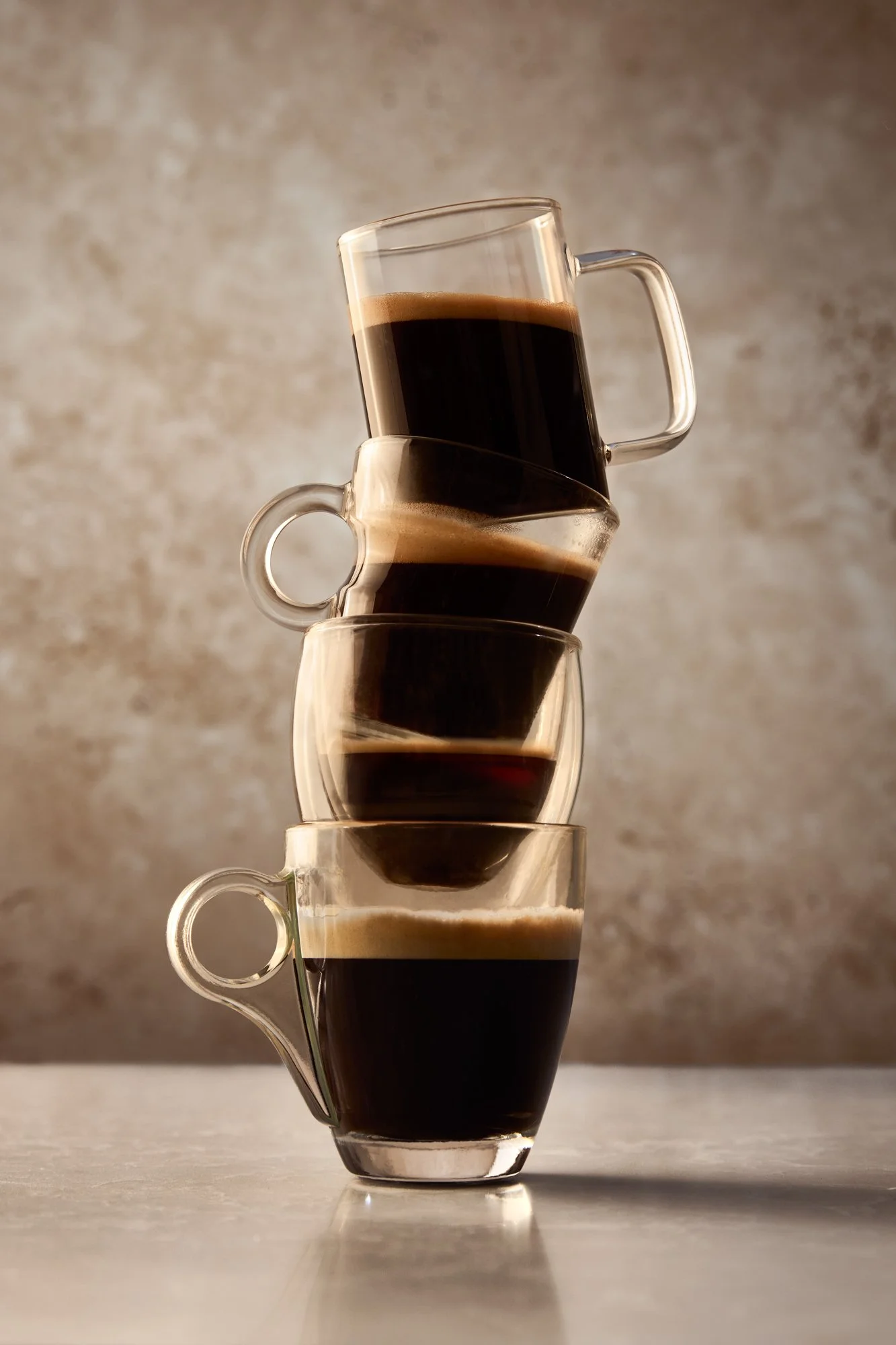 A stack of four glass cups filled with black coffee, arranged with three cups upright and one cup tilted on top, on a light-colored surface against a textured wall background.