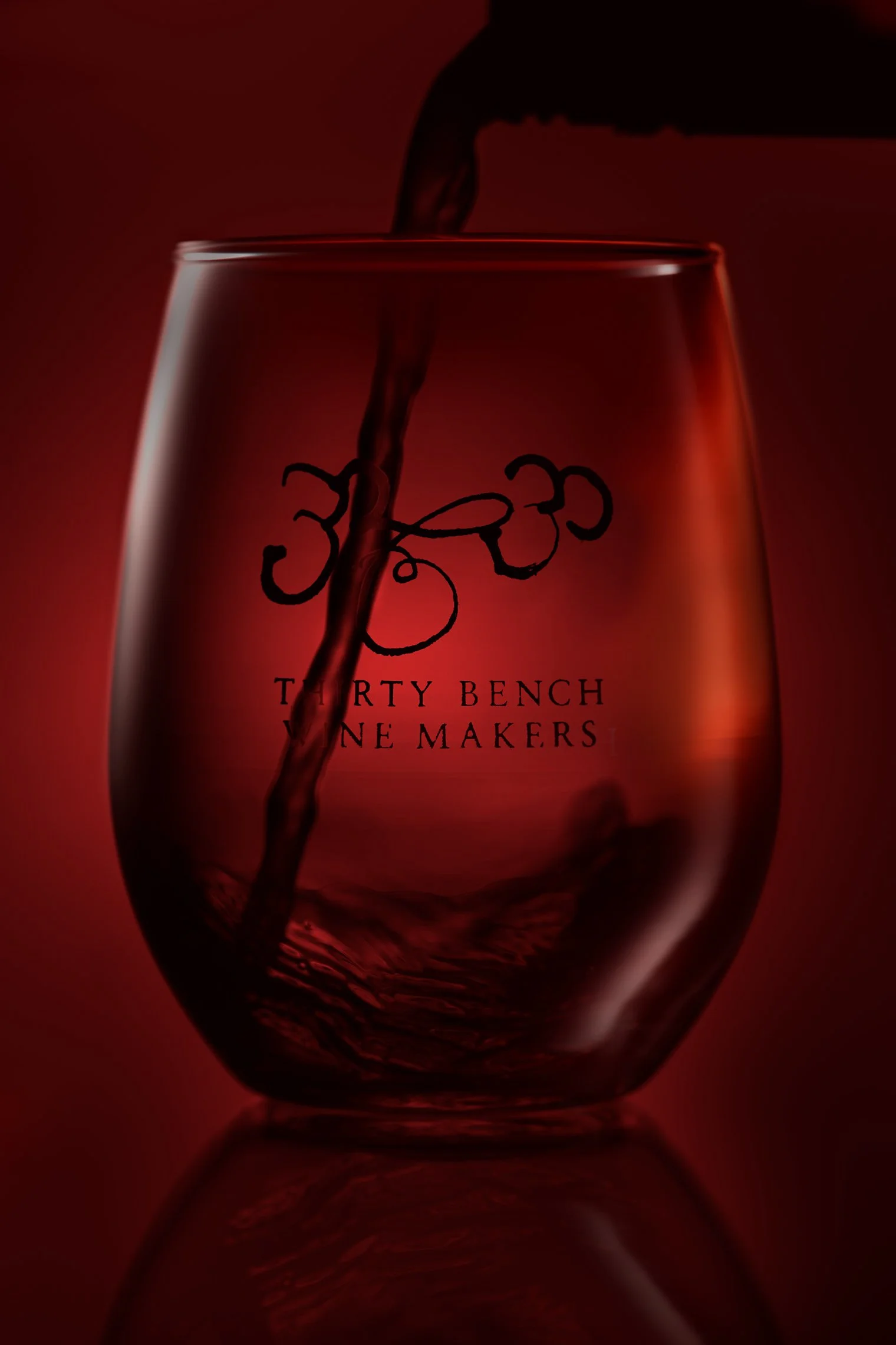 A glass of red wine being poured, with a logo and text on the glass that reads 'Thirty Bench Wine Makers'.