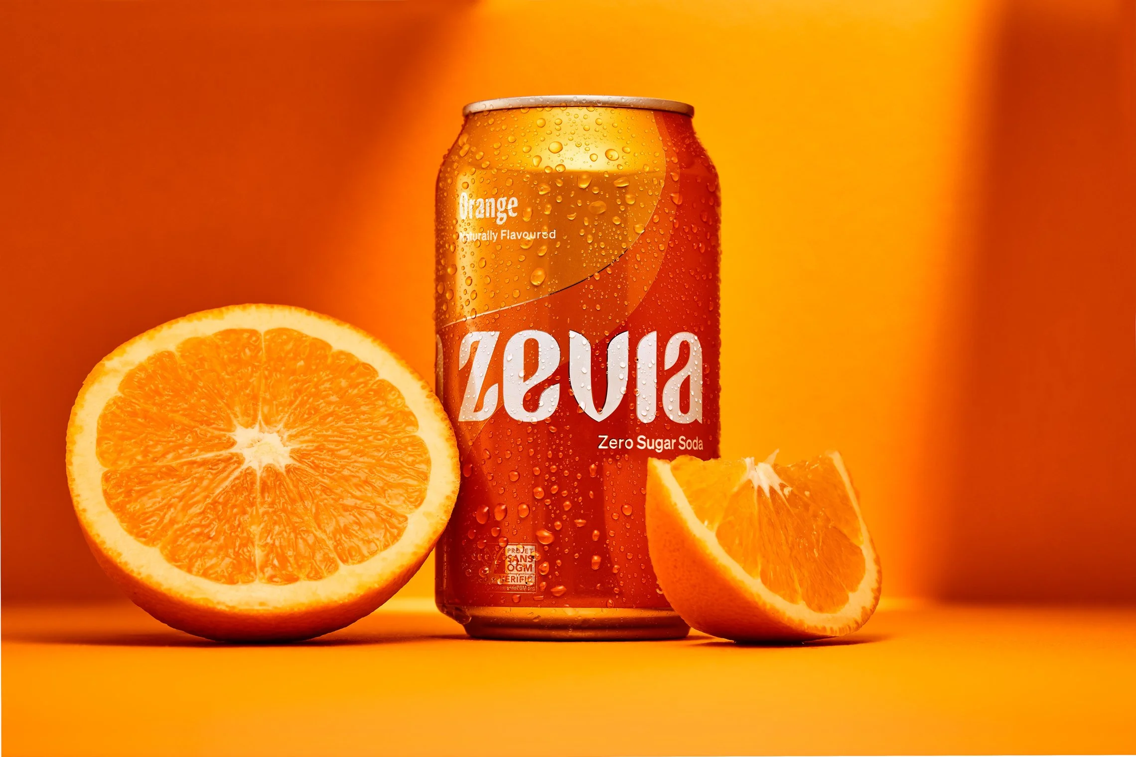 A can of Zevia orange flavored zero sugar soda next to a halved orange and an orange wedge on an orange background.