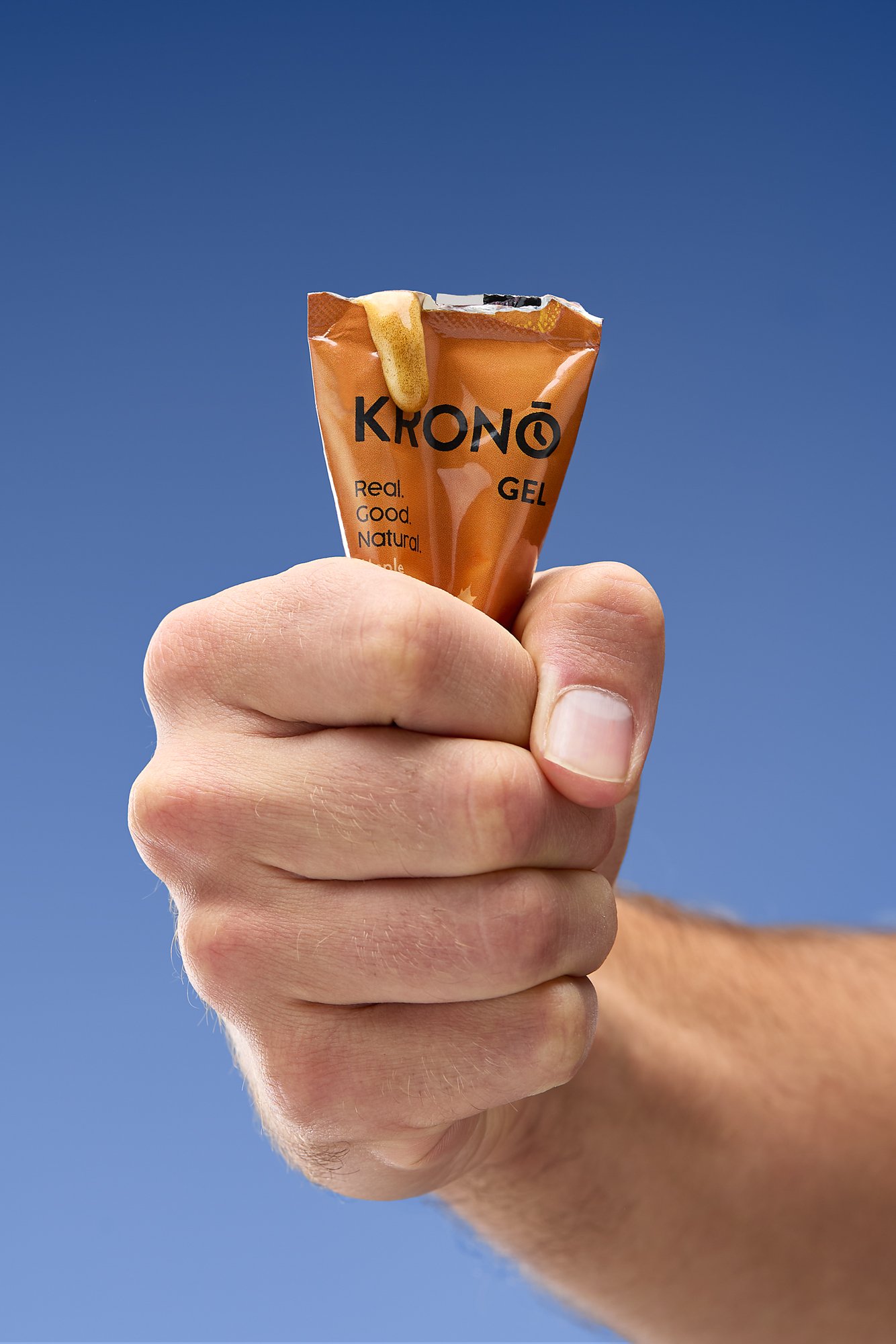 A person's hand holding a small orange packet labeled 'KRONO GEL' against a clear blue sky, with some gel spilling out of the top.