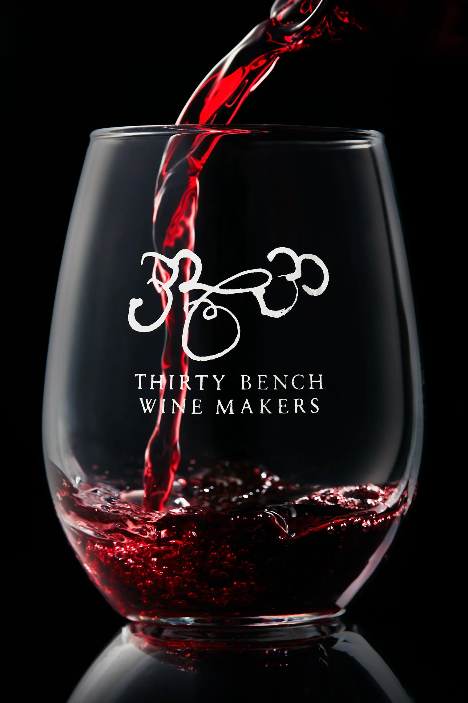 Red wine being poured into a transparent wine glass with the logo and name 'Thirty Bench Wine Makers' on the glass, against a black background.