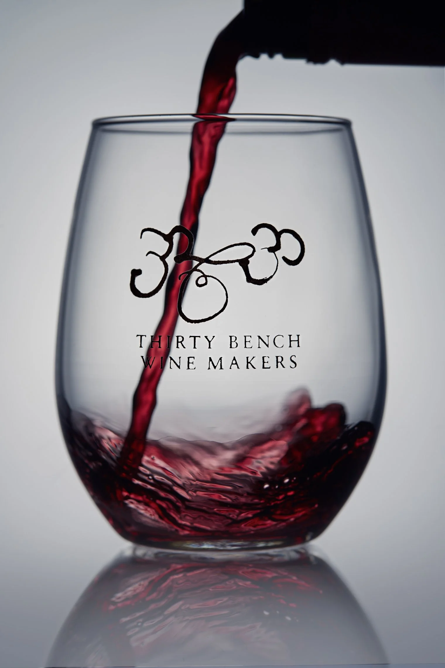 Red wine being poured into a clear glass with the logo and text 'Thirty Bench Wine Makers' on it.