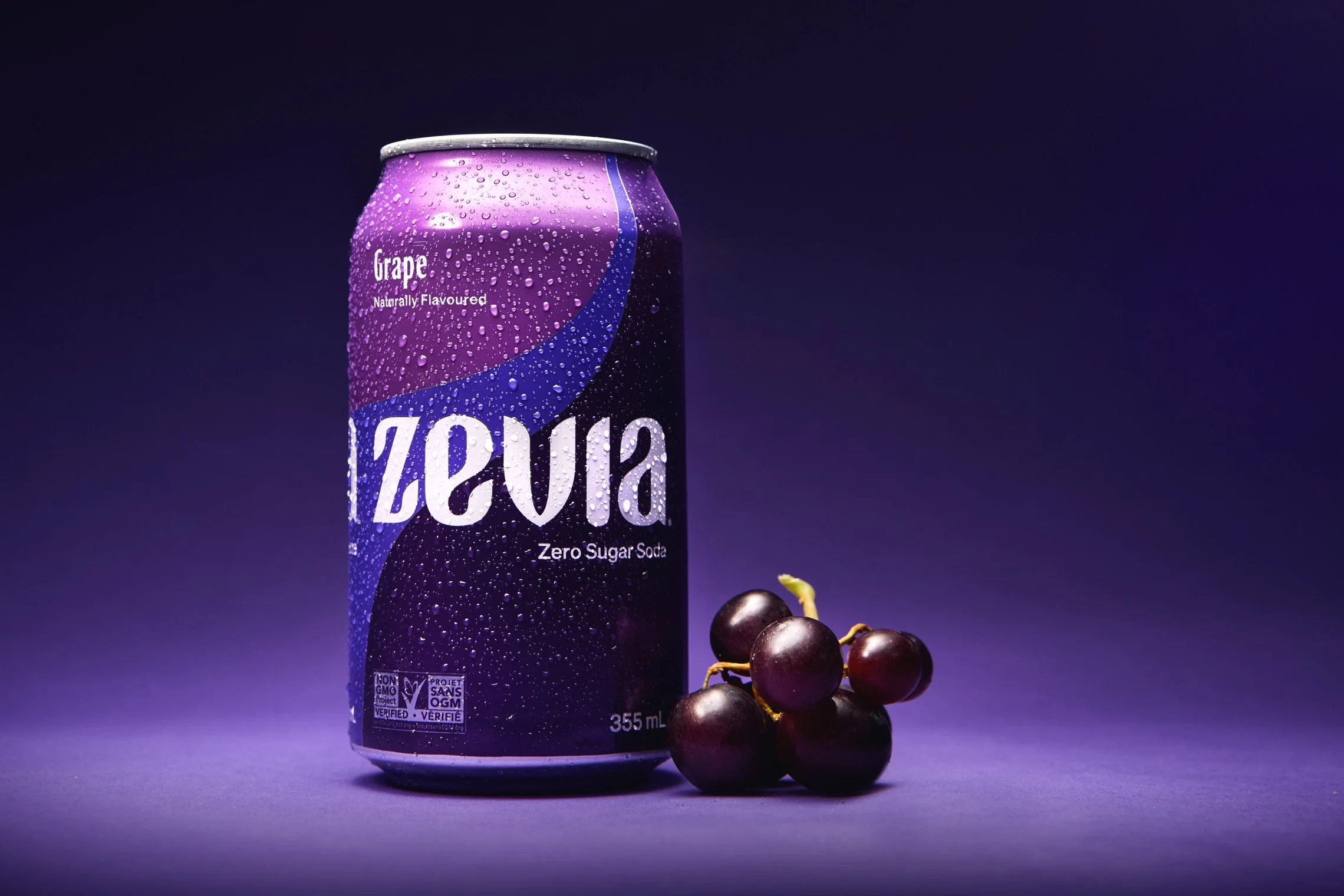 A can of Zevia grape zero sugar soda with a bunch of black grapes beside it, set against a dark purple background.