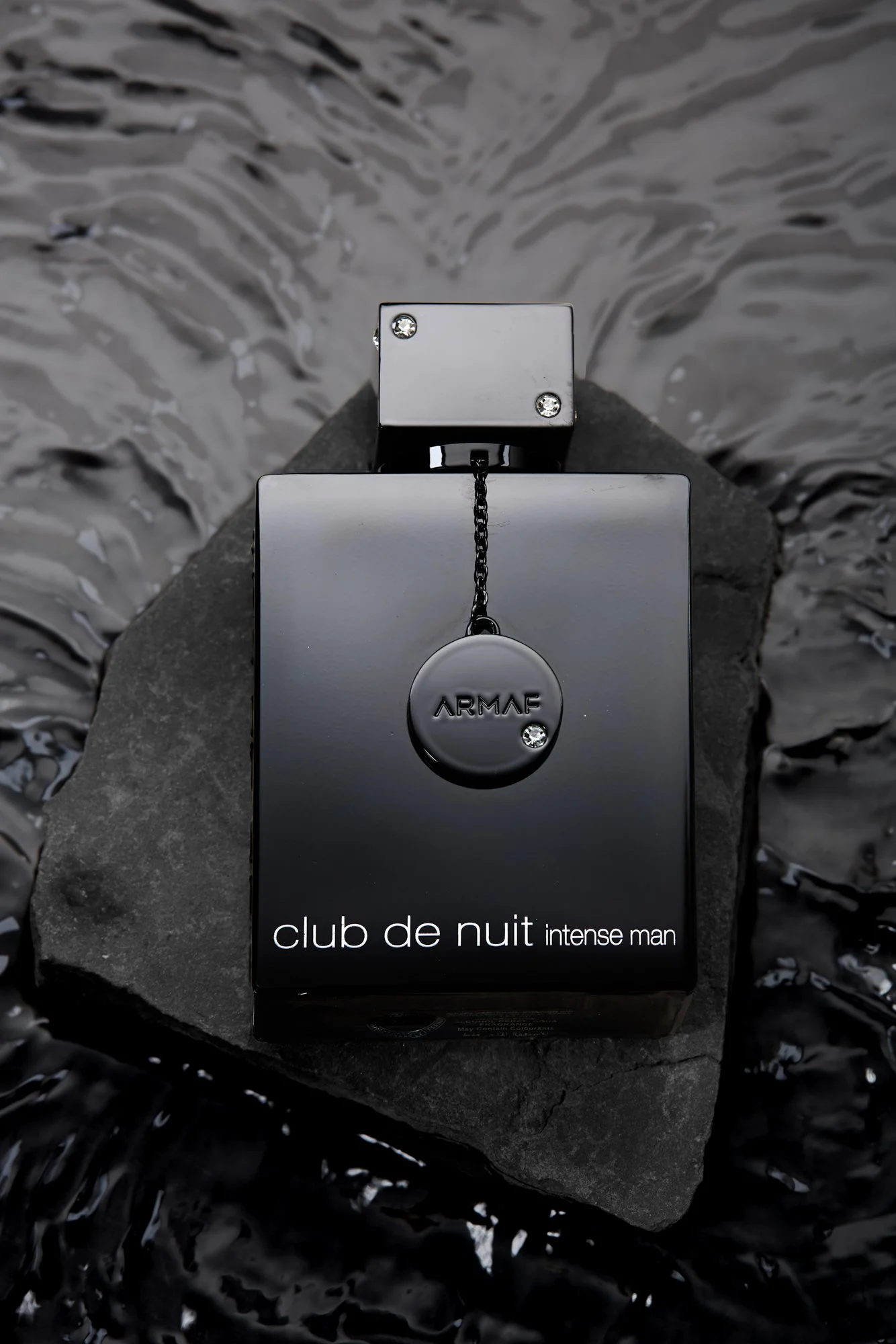 Black perfume bottle labeled 'club de nuit intense man' with a spherical cap that says 'ARMAF,' placed on a gray rock near water.