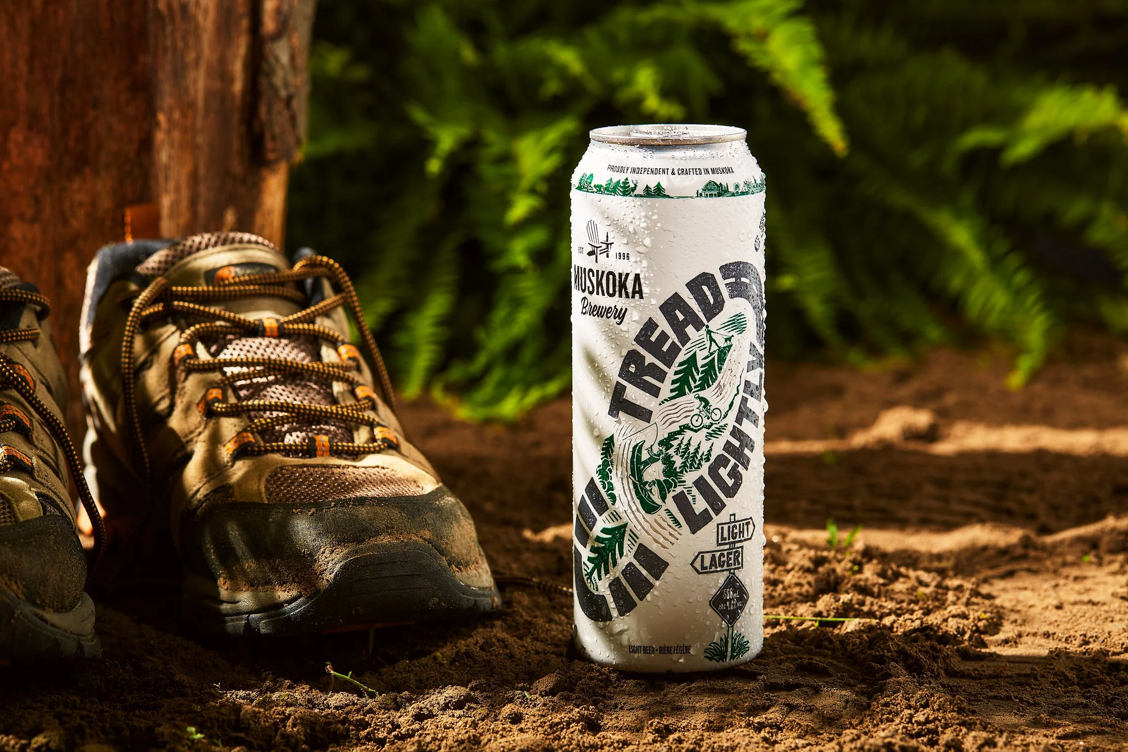 A can of Trailhead Brewing Light Lager beer stands on dirt next to a pair of hiking shoes and green foliage in the background.